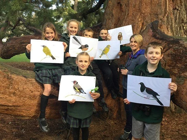 Group of six children and one adult outdoors, holding bird illustrations in front of a large tree.