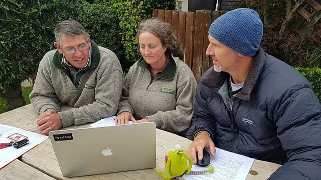 Three people sitting at a picnic table outdoors, looking at a laptop. Two are men and one is a woman, and all are focused on the laptop screen, with notebooks and papers around them.