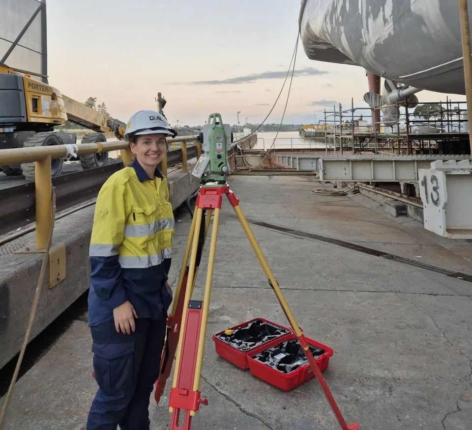 A women wearing a hard hat and overalls standing at a shipping dock next to a theodolite.
