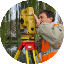 A person operating a yellow surveying instrument outdoors near water and trees.