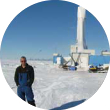 A man standing outdoors in a snowy environment near industrial equipment under a blue sky.