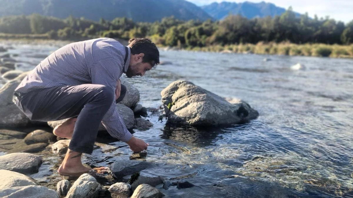 A man crouched by a river reaching into the water.