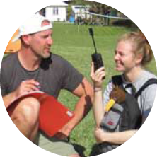 A man and a woman sitting outdoors on grass, smiling and talking. The man is holding a walkie-talkie, and the woman has a backpack. It appears to be a sunny day.