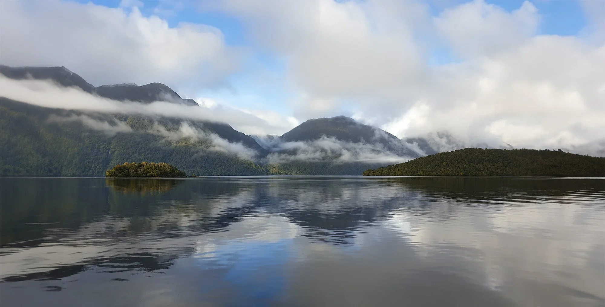 Calm lake with reflections, surrounded by forested mountains partly covered with clouds and mist, under partly cloudy sky.