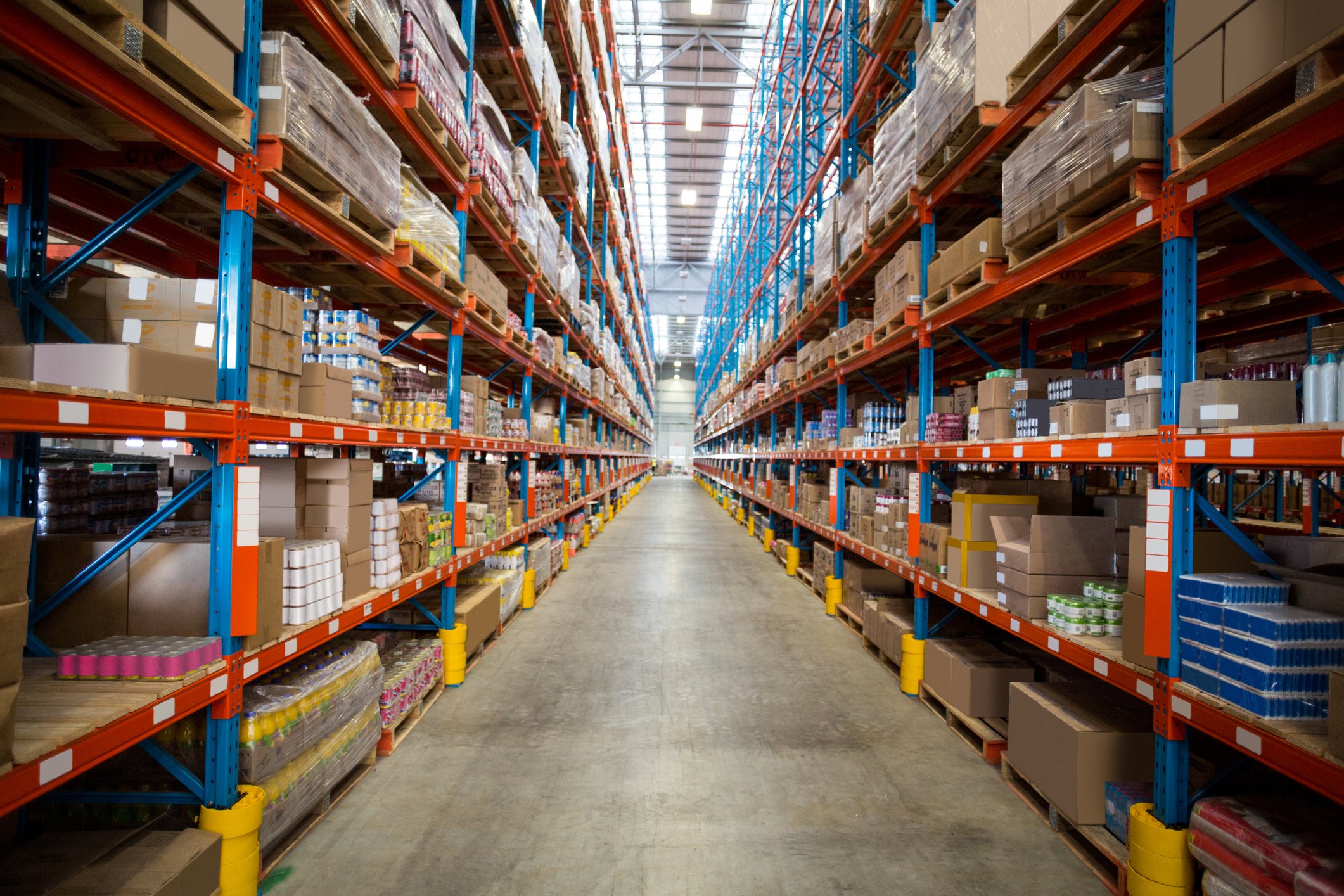 A large warehouse aisle with tall shelves on either side filled with cardboard boxes and packages. The warehouse has a high ceiling with skylights, and the floor is clear and empty, extending into the distance.