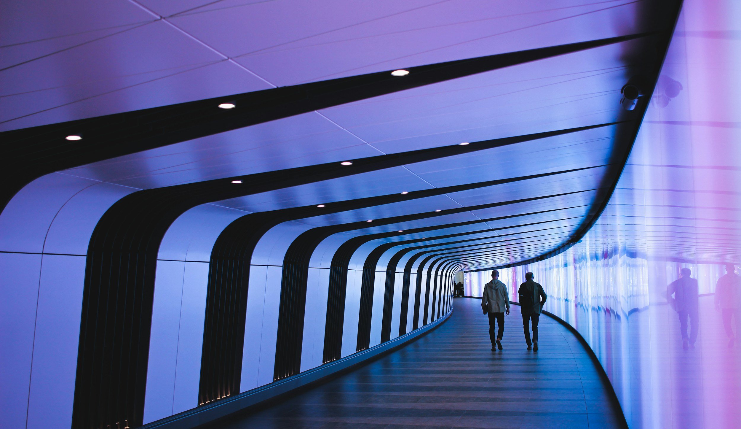Two people walking through a modern, curved corridor with a blue and purple illuminated ceiling.