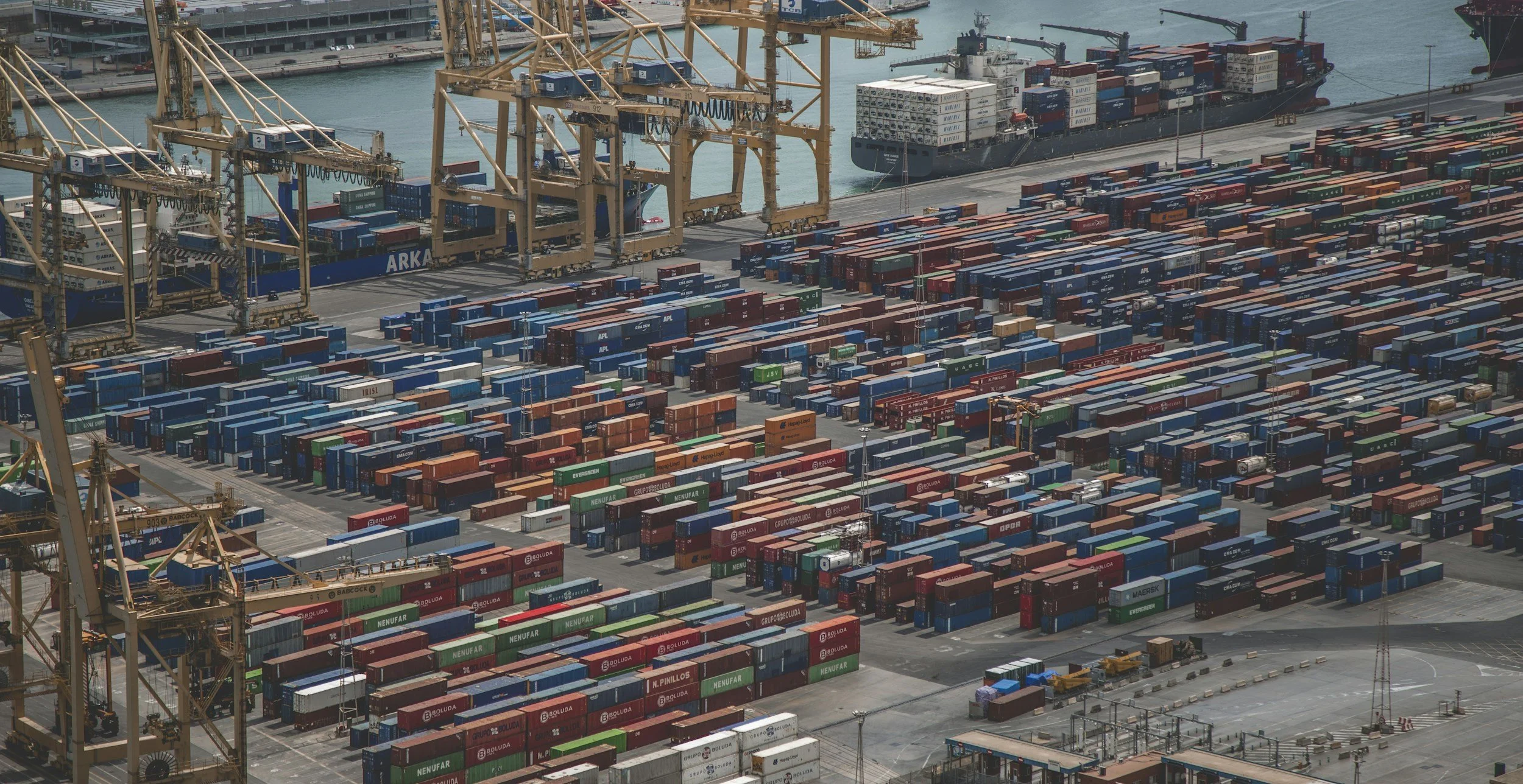 View of a busy shipping port with numerous cargo containers stacked in rows, several large cranes for loading and unloading ships, and a cargo ship docked in the background.