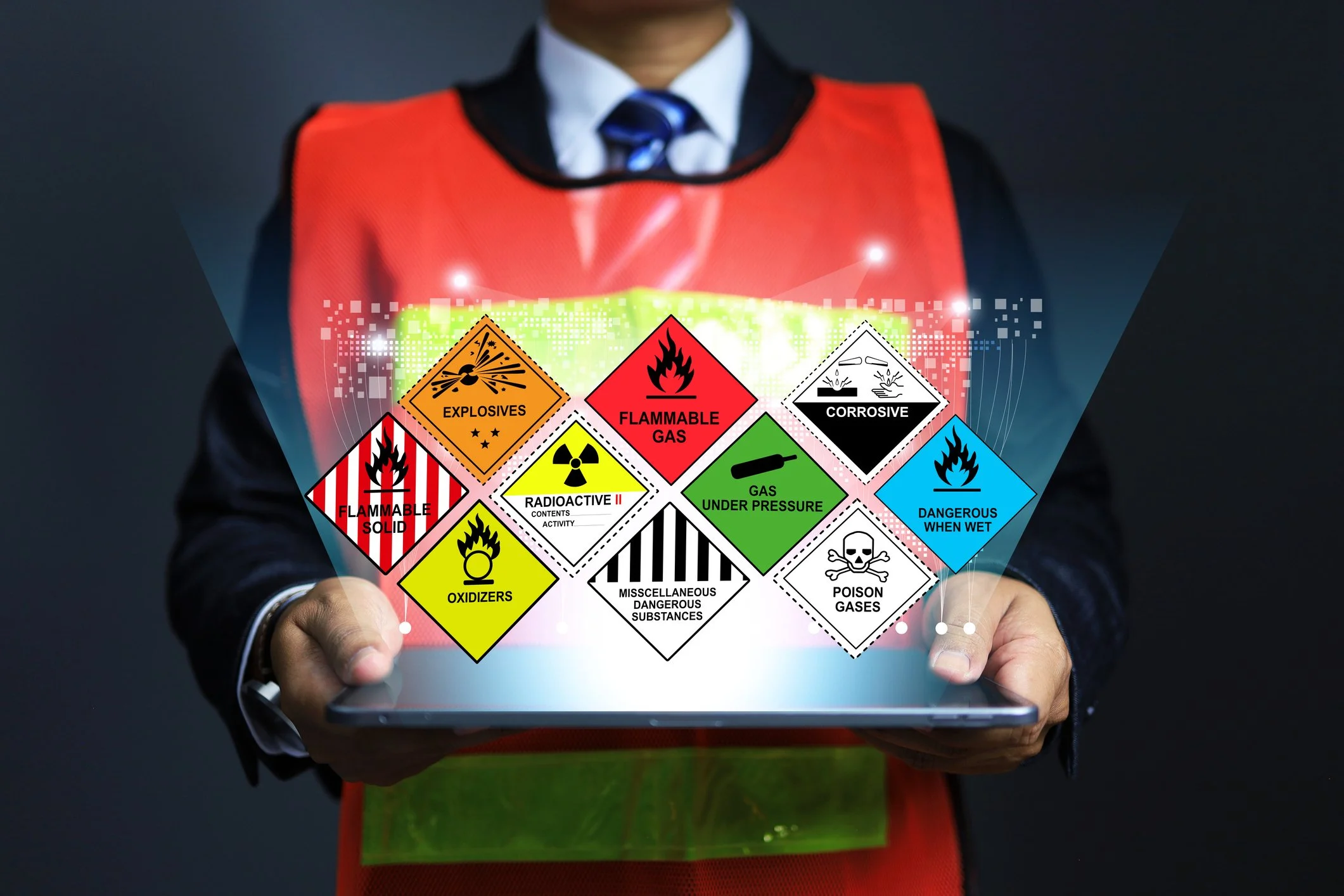 Person wearing a black suit and a red safety vest holding a tablet with various hazardous material warning labels floating above it, including flammable, explosive, radioactive, corrosive, toxic, and other hazard symbols.