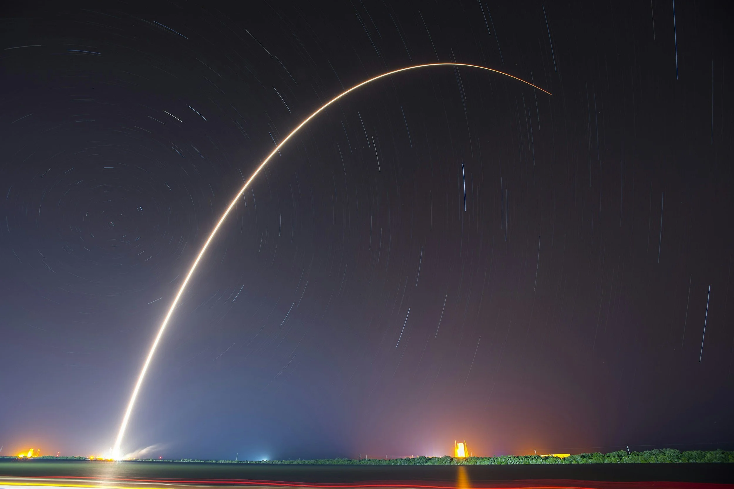 A long exposure photo of a rocket launch showing a bright trail against the night sky, with star trails, a distant planet or star, and some illuminated structures on the horizon.