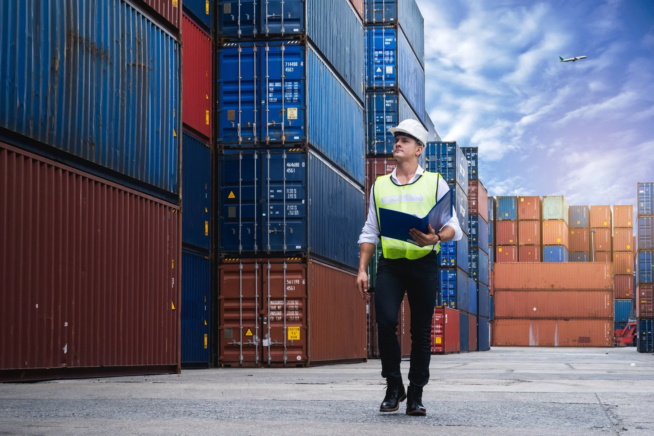 A man in a white safety helmet and reflective vest walking through a shipping yard with stacked cargo containers, with an airplane flying in the cloudy sky above.