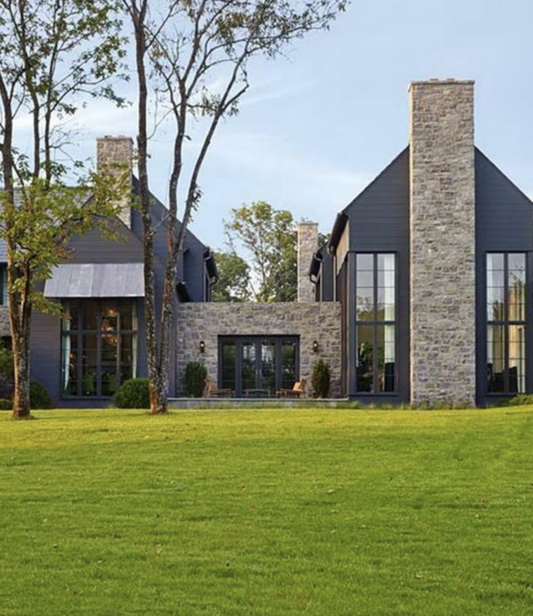 Modern house with stone and glass facade, large windows, lawn in foreground, and trees surrounding the property.