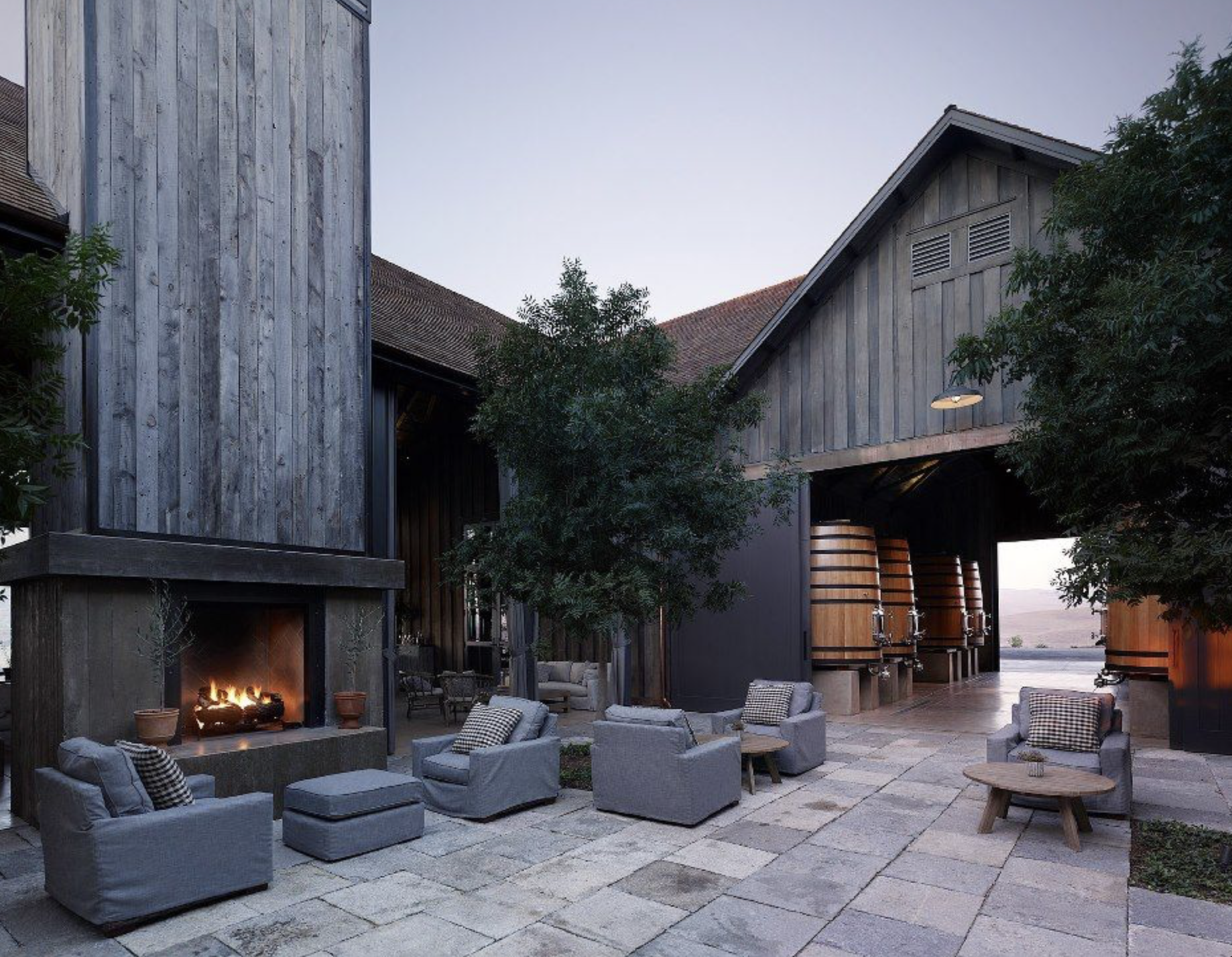 Outdoor patio with fireplace, gray chairs, and large wooden barrels inside a rustic building with trees nearby.