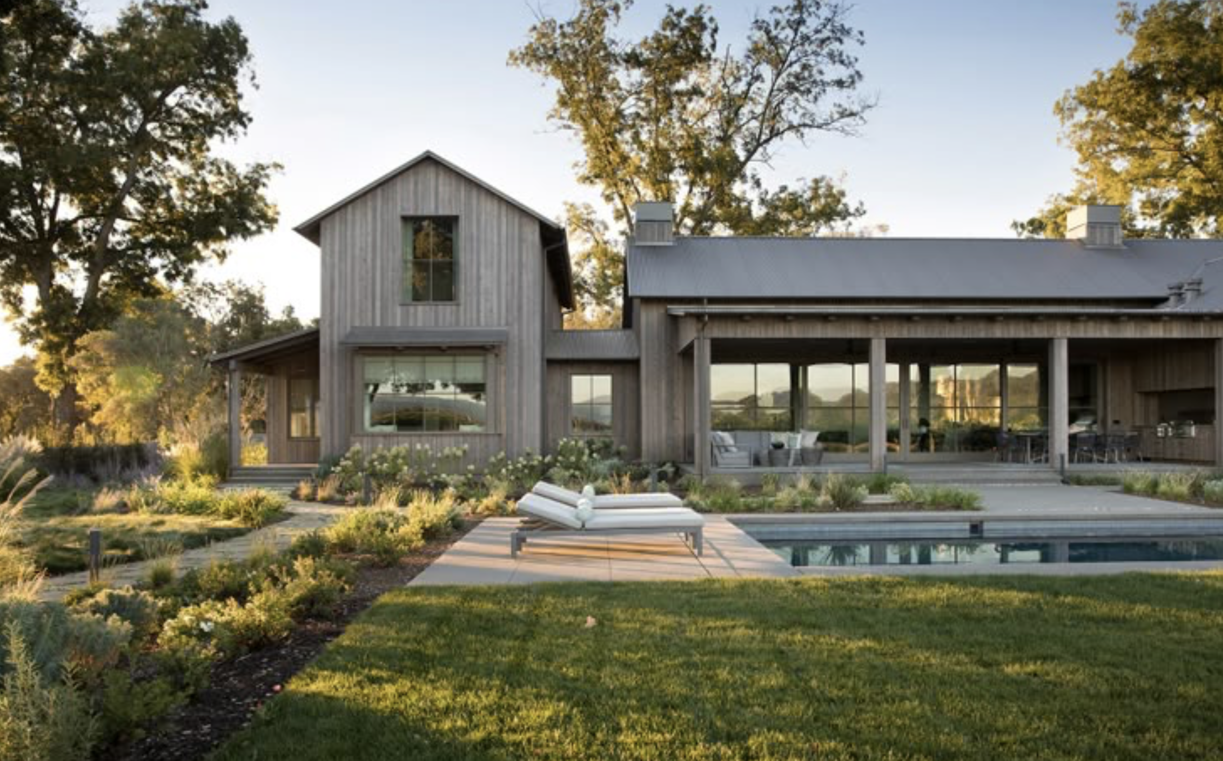 Modern farmhouse with wooden exterior, large windows, outdoor patio, swimming pool, and garden, surrounded by trees.