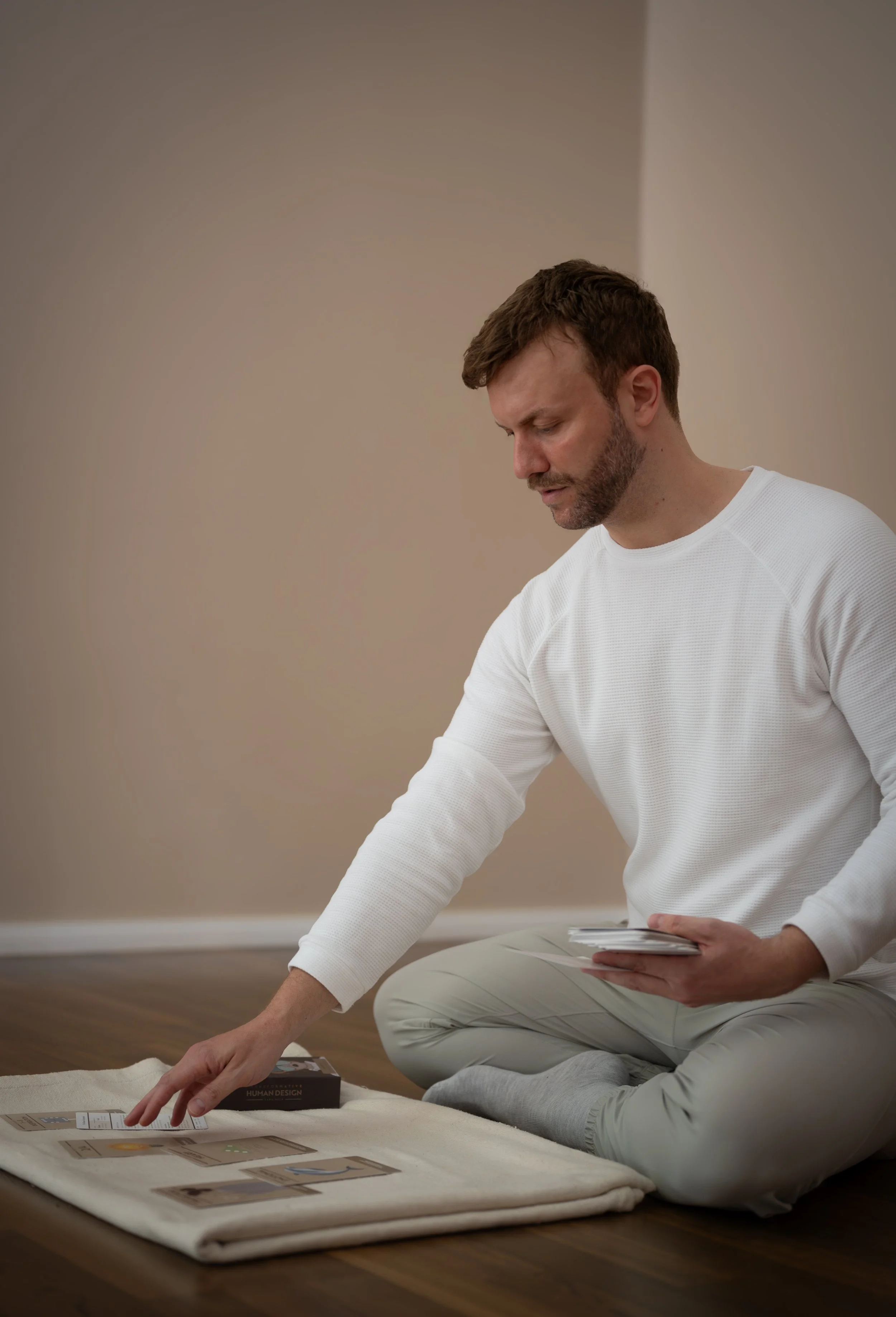 A man with a beard, wearing a white long-sleeve shirt, is kneeling on the floor, looking at design cards laid out on a table. He is holding a stack of cards in his left hand, and his right hand is reaching towards the cards on the table. The background is a plain wall, and the floor is wood.