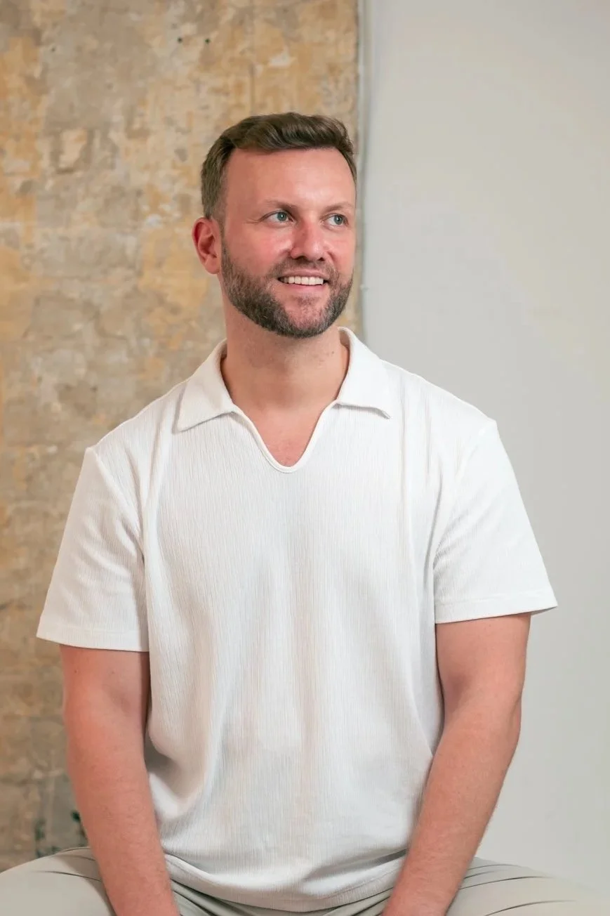 A smiling man with brown hair and a beard, wearing a white short-sleeved collared shirt, sitting against a brick wall and off-white wall background.