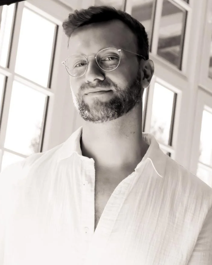 A man with glasses and a beard, wearing a light-colored shirt, standing indoors near large windows with natural light in the background.