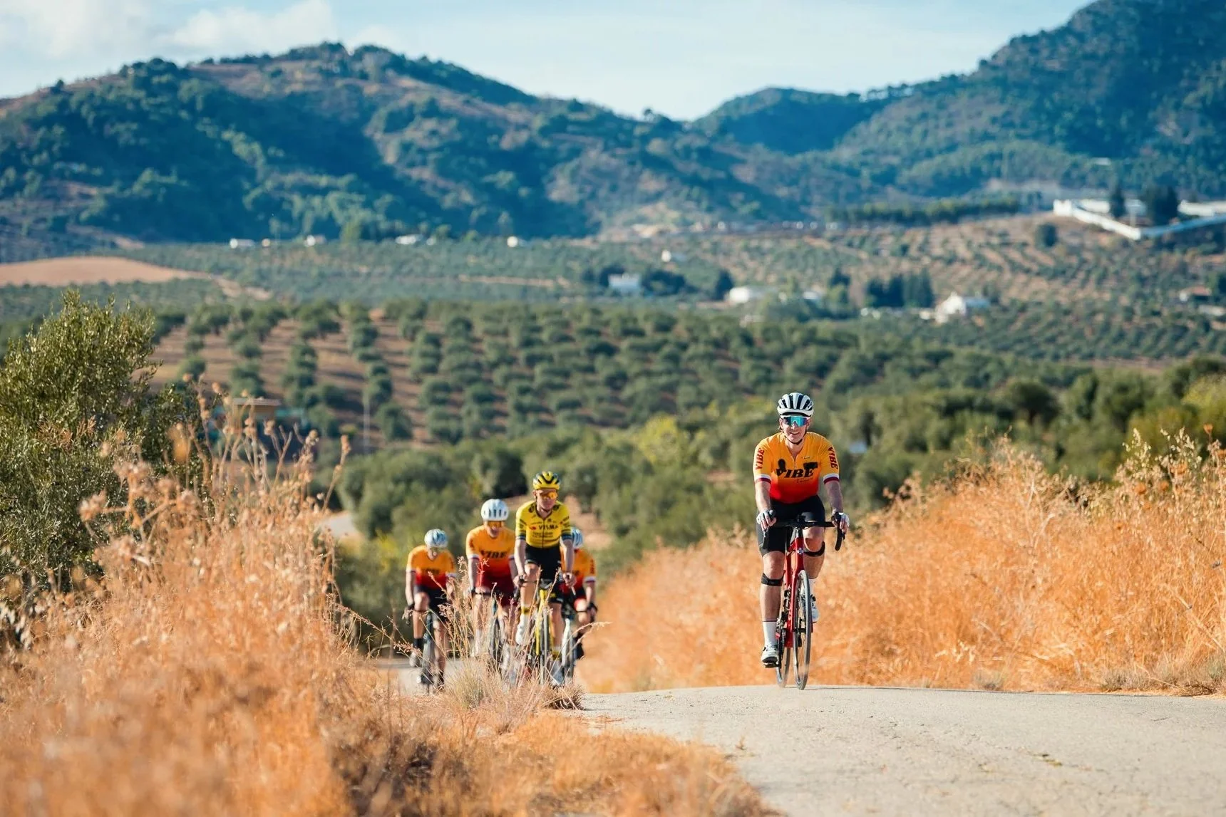 Cyclists riding with Sepp Kuss on a rural road with hills and mountains in the background.