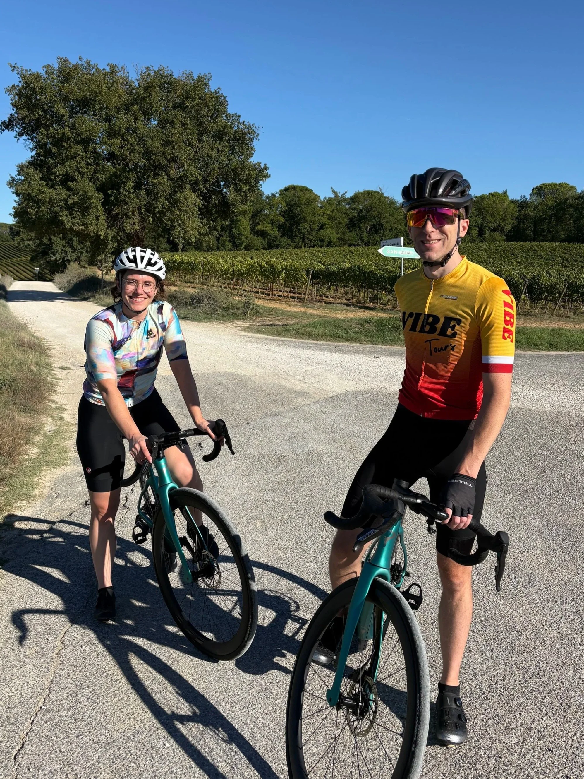 Cyclists wearing helmets and sunglasses riding a bicycle on a mountain road with a gravel road and vineyard in the background.