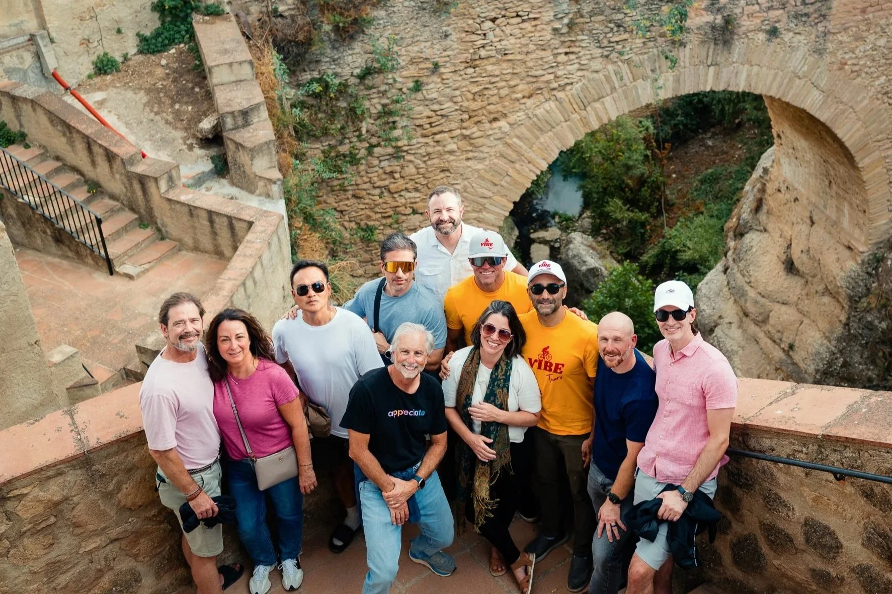 A group of tourists poses for a photo in front of an arched stone bridge spanning the gorge in Ronda, Spain.