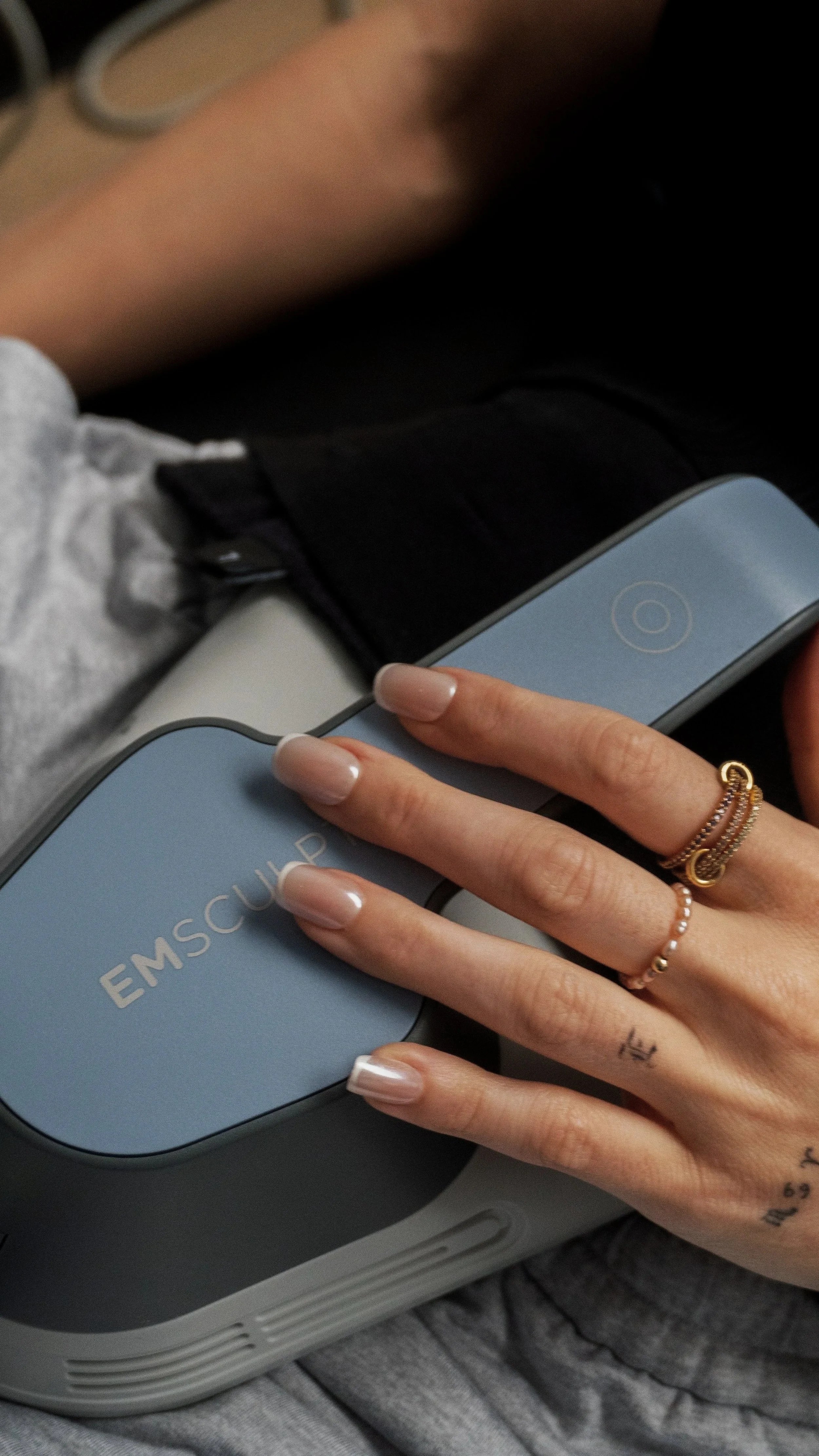 A person's hand resting on a medical device labeled 'EMS Coupe,' with the words in gold, in a relaxed setting with casual clothing and jewelry.