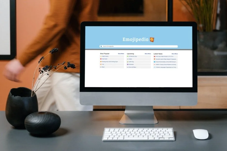 Desktop computer screen on a desk next to a vase. Emojipedia.com is on the screen.