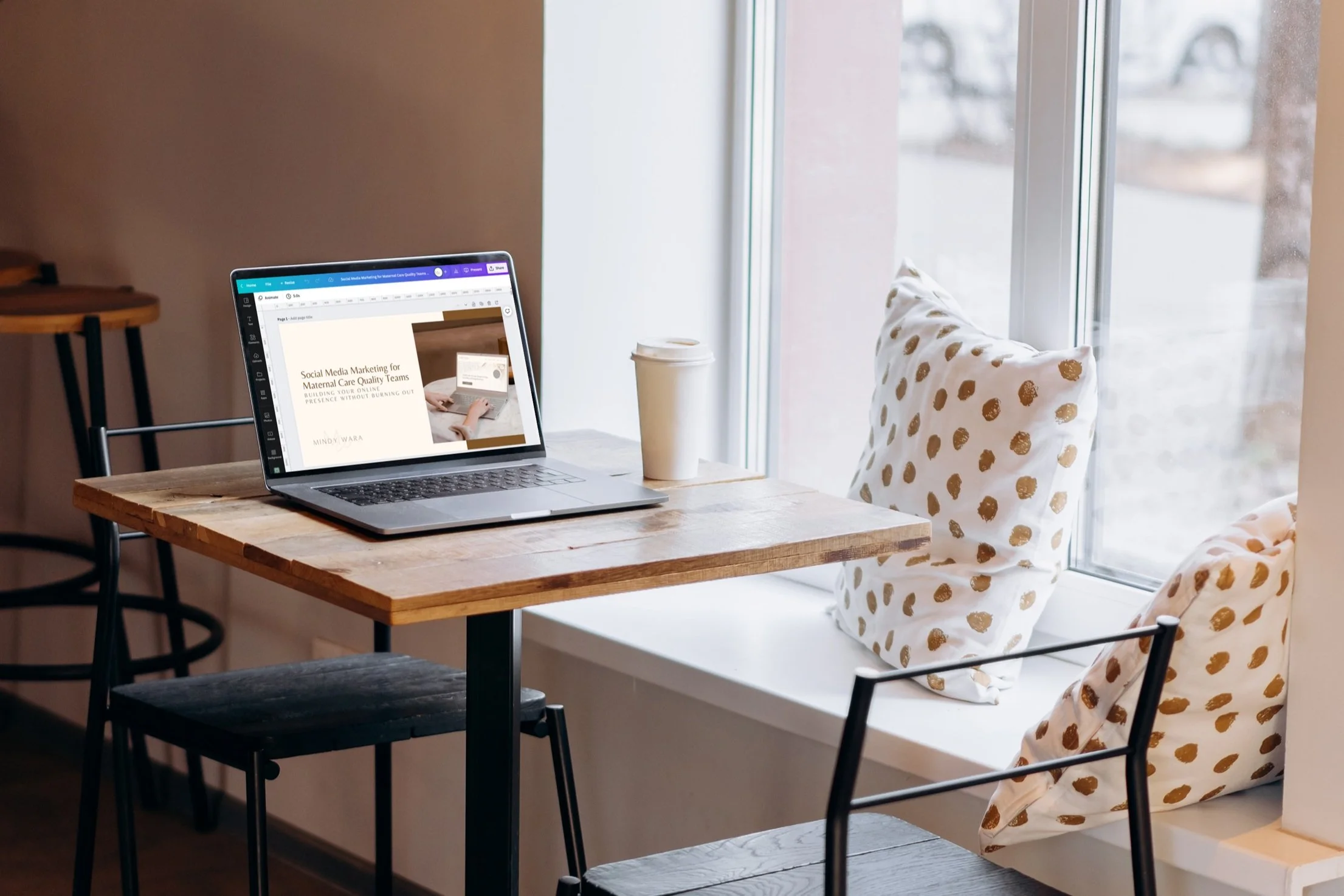 Laptop sitting next to a paper coffee cup on a cafe table next to a window. Canva.com is on the screen.