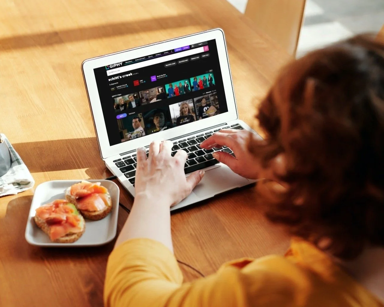 Red-headed person facing a small laptop while sitting at the dinner table with a sandwich. Giphy.com is on the screen.