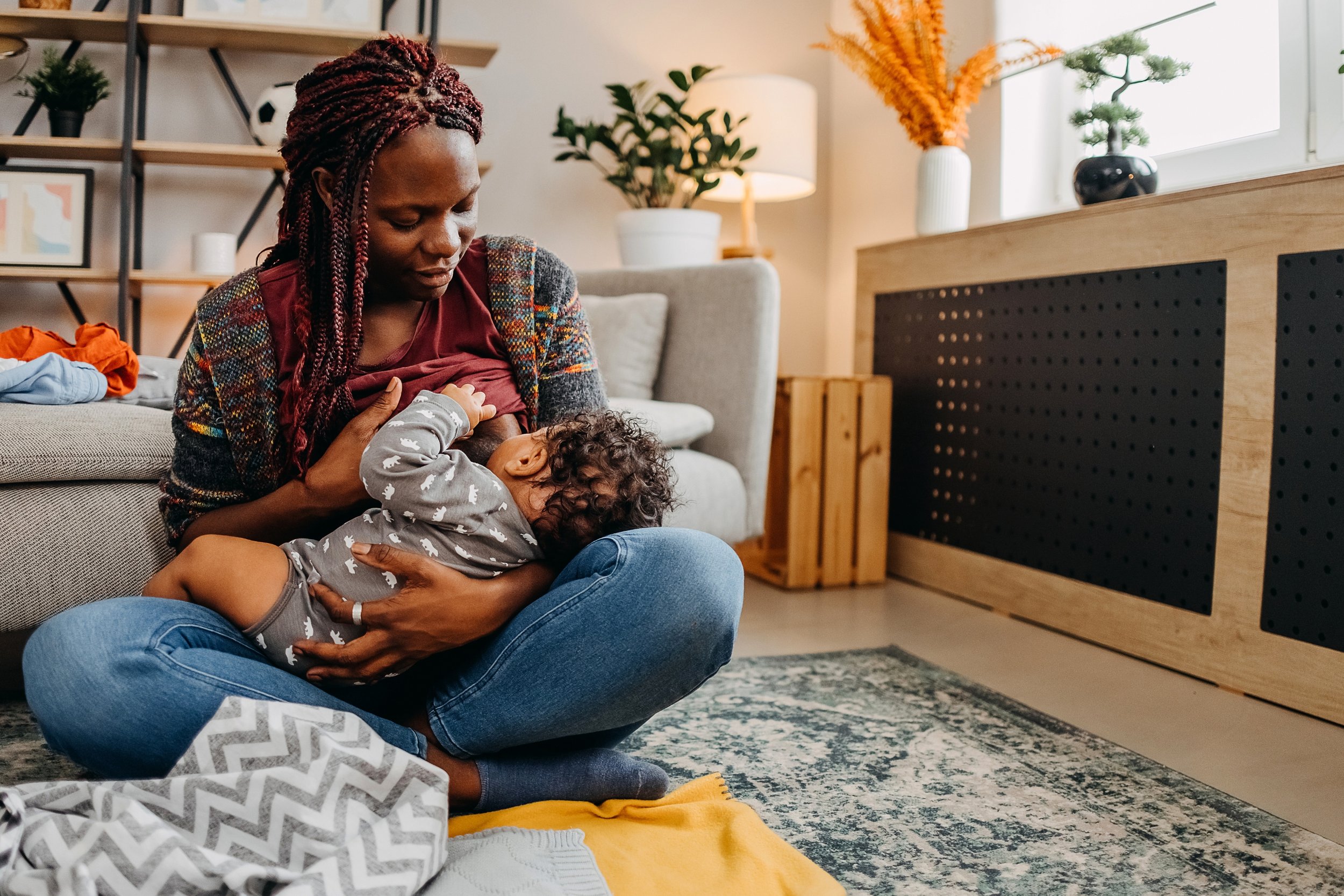A parent sits cross-legged on a rug in a cozy living room, breastfeeding a toddler. The parent looks down gently at the child, surrounded by soft blankets.