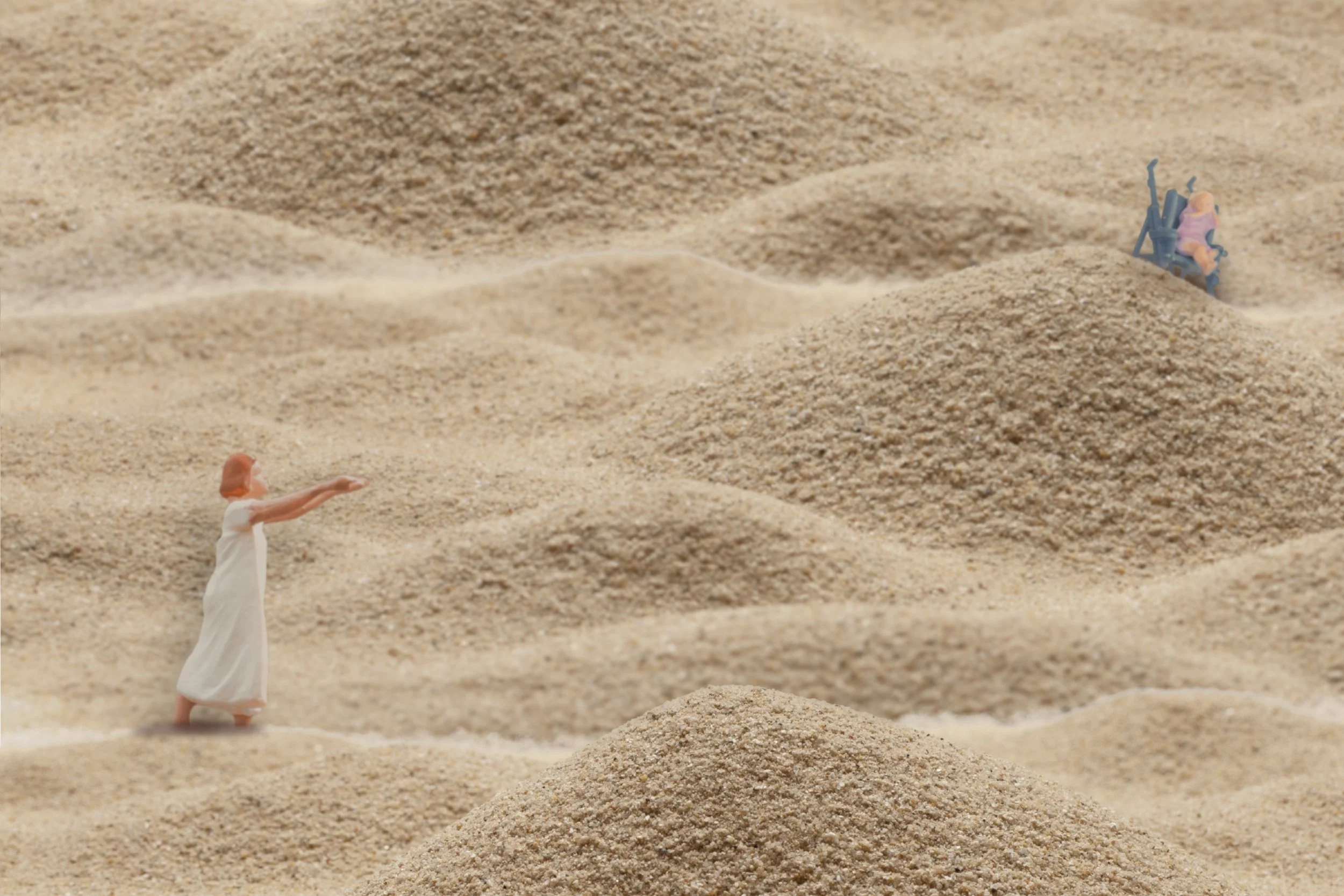 A miniature of a woman in a hospital gown stands in a sandy landscape, arms reaching toward a distant stroller with a small figure of an infant, separated by mounds of sand.
