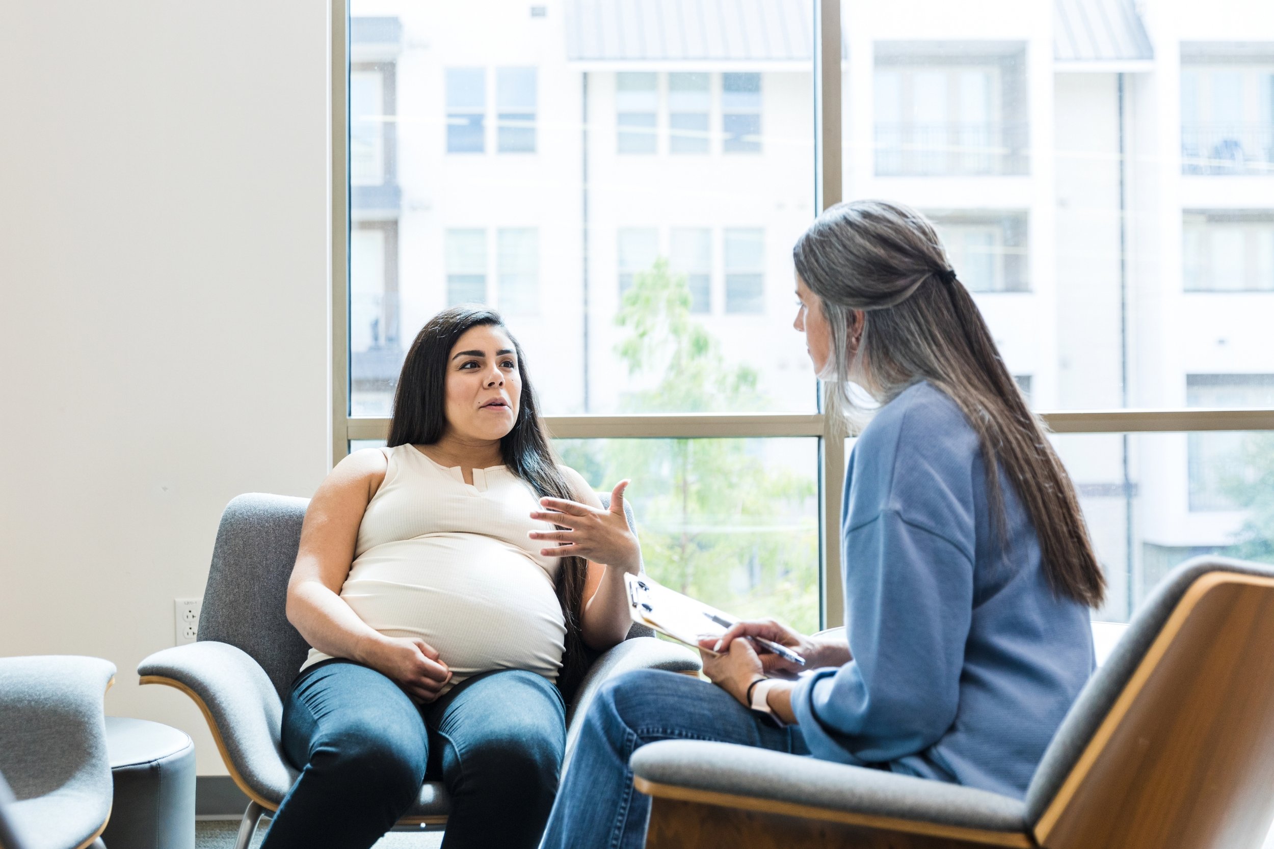 A pregnant client sits in conversation with a therapist in a modern, sunlit counseling office. Both appear engaged in discussion, suggesting a supportive therapeutic environment.