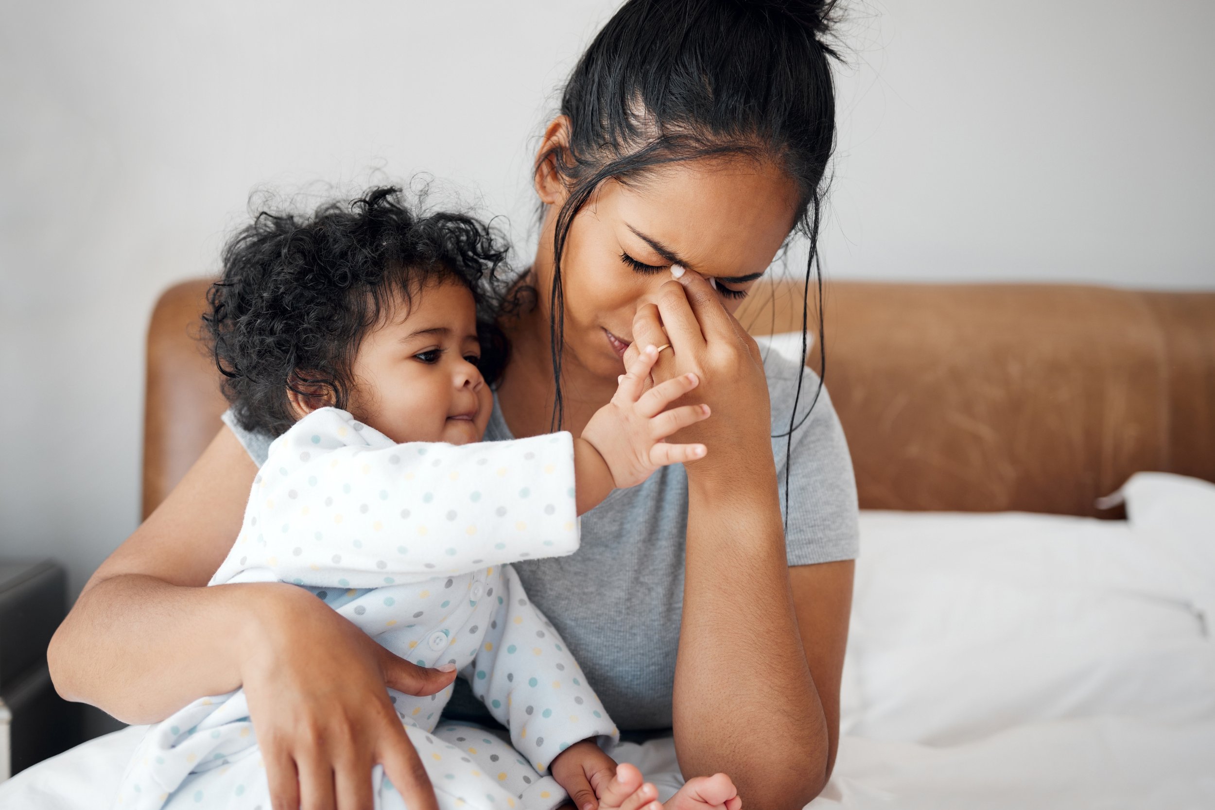 A mother sits on the edge of a bed, holding her child while rubbing her eyes in frustration and fatigue. The baby reaches toward her face, unaware of her emotional state. The image captures a moment of quiet overwhelm.
