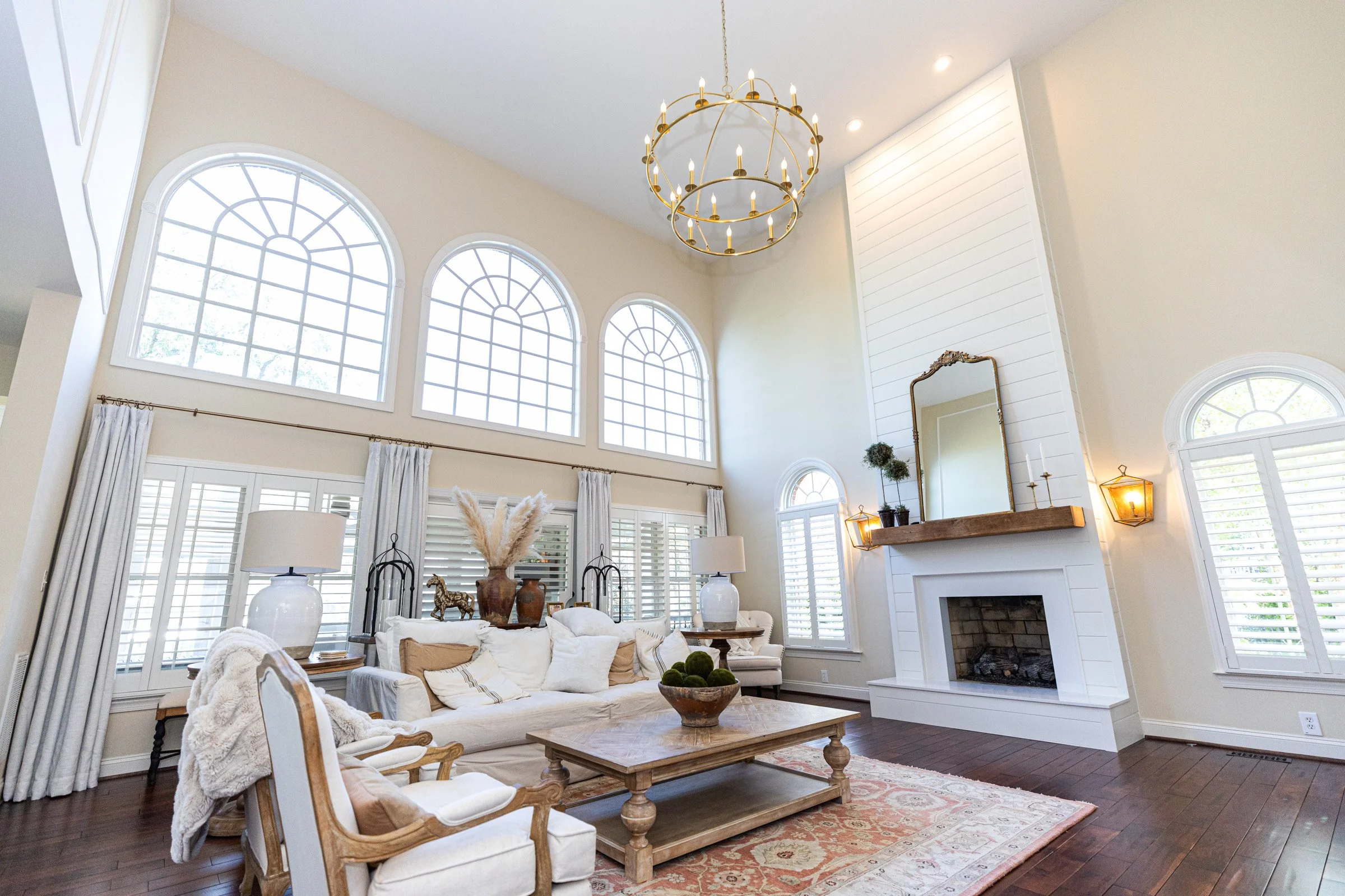 A spacious living room with large arched windows, a white fireplace, a chandelier, and a cream-colored sofa with pillows.