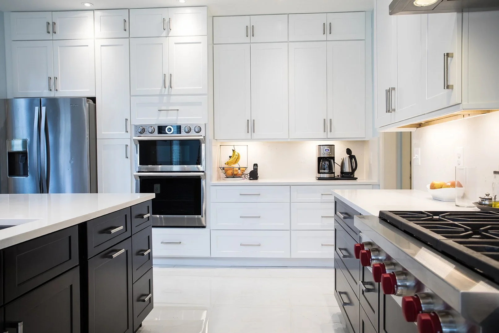 Kitchen with white upper cabinets, black lower cabinets, stainless steel refrigerator, oven, coffee machine, toaster, bananas in a basket, fruit on the counter, and a gas stove with red knobs.