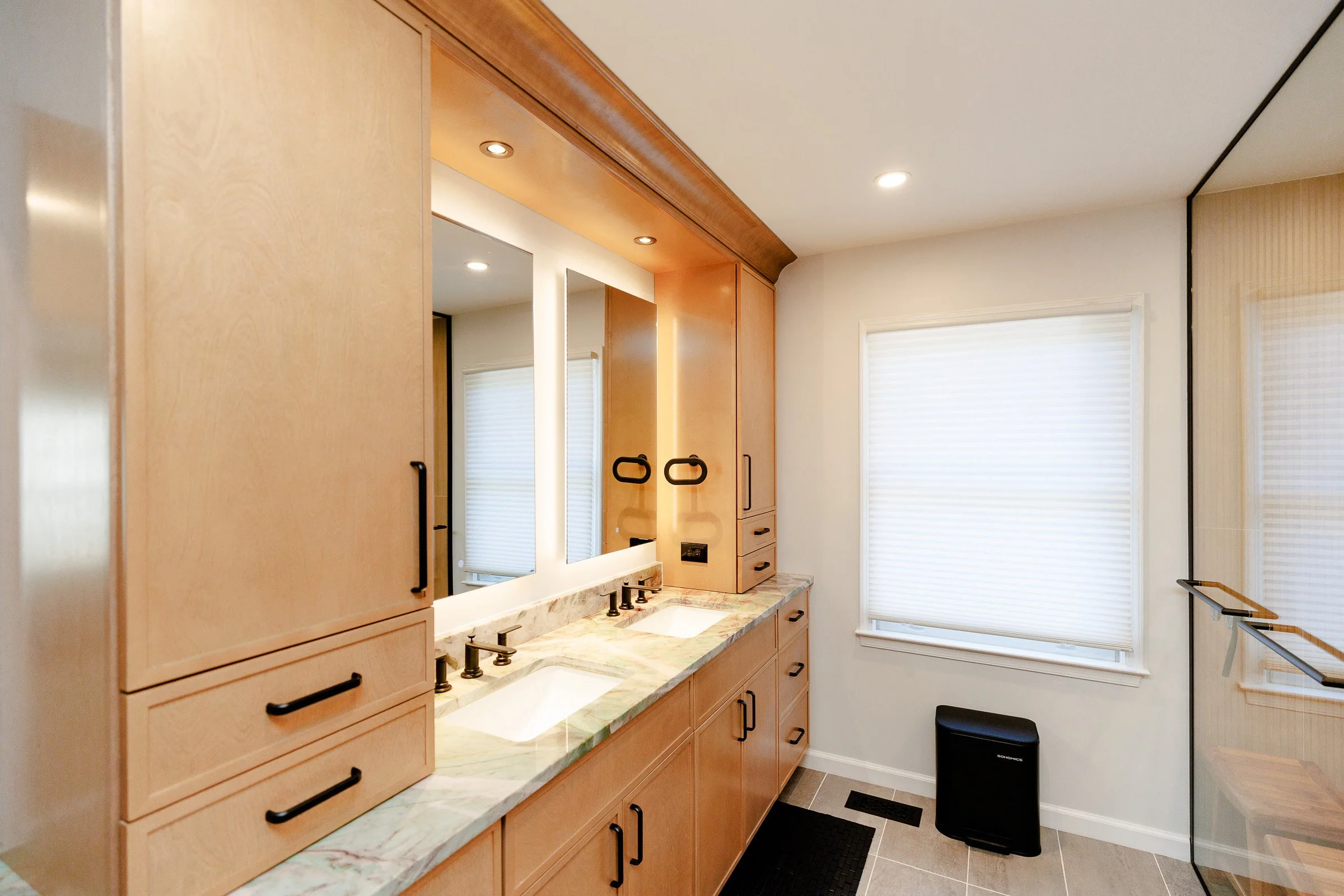 Bathroom with wooden cabinetry, marble countertop, double sinks, white window blinds, beige walls, small black trash bin, and black towel rack.