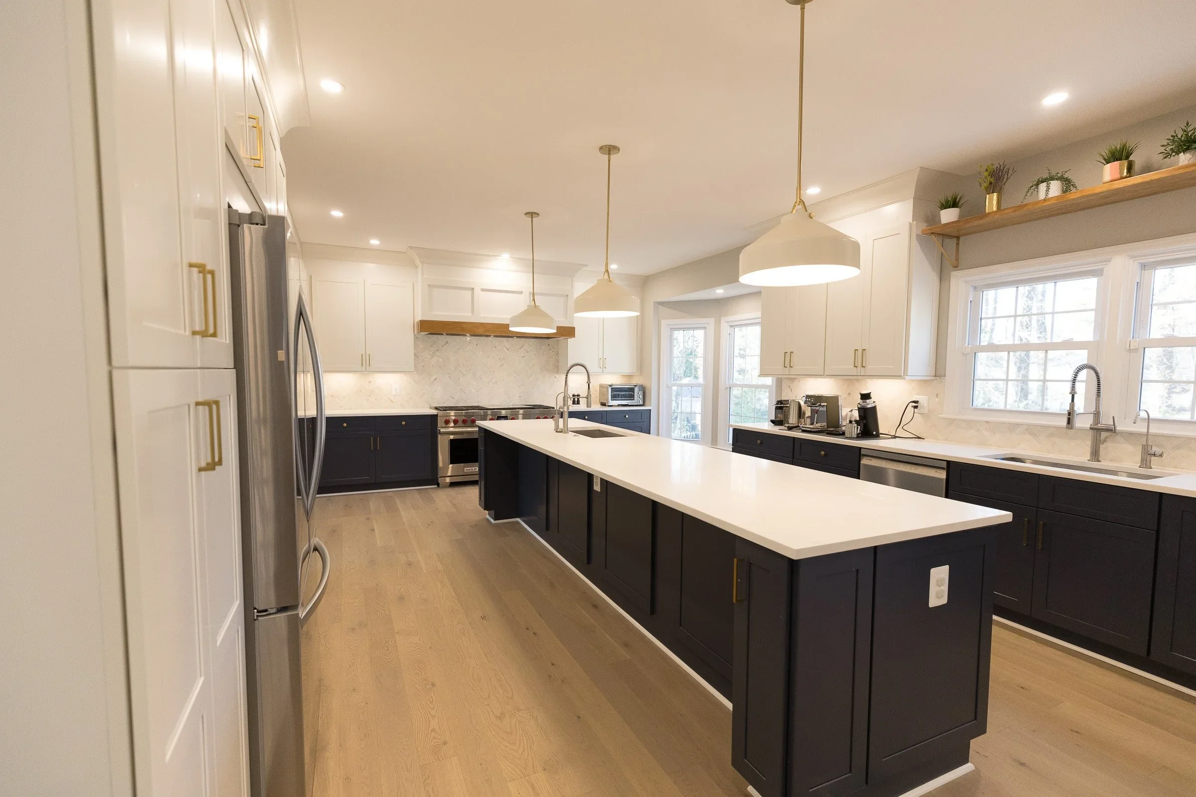 Modern kitchen with white upper cabinets, dark lower cabinets, a white island, pendant lights, appliances, and large windows with natural light.