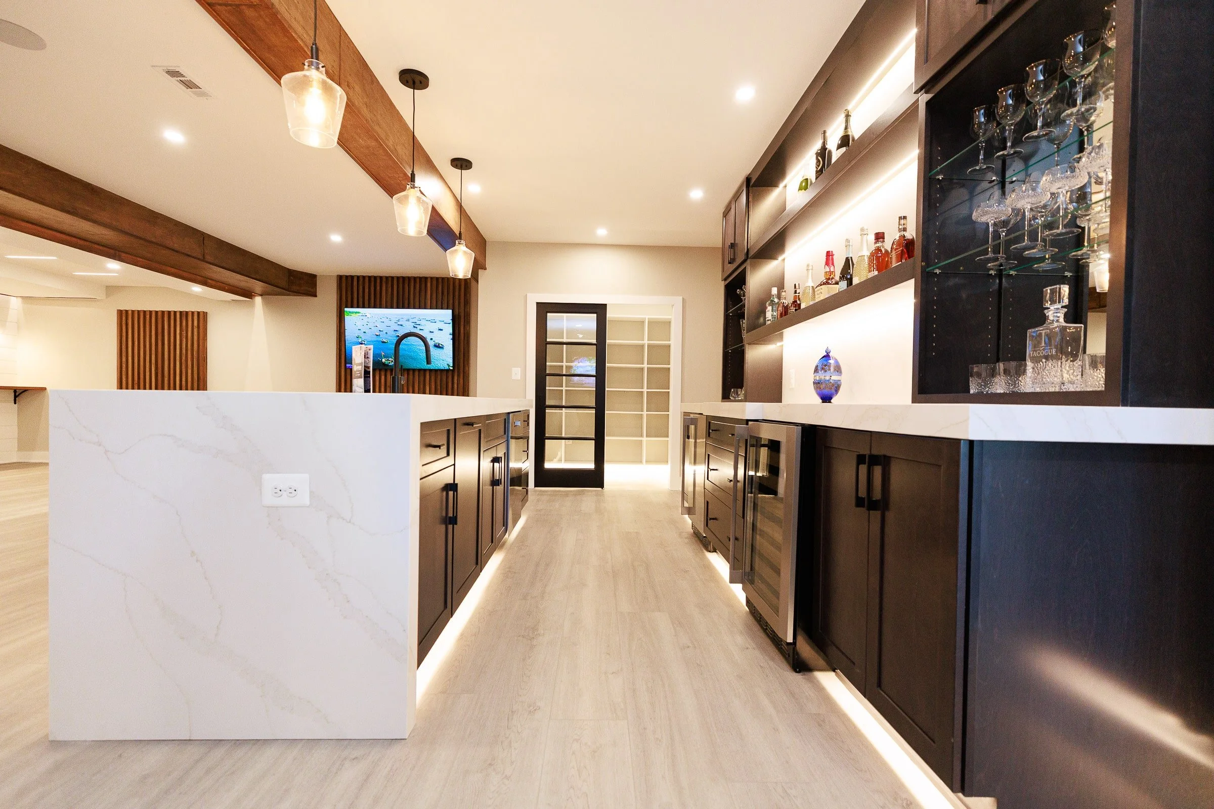 Modern kitchen with dark cabinets, white countertops, and under-cabinet lighting. There is a bar area with glassware and bottles, pendant lights hanging from a wooden beam, and a sliding door leading to a room with built-in shelving.