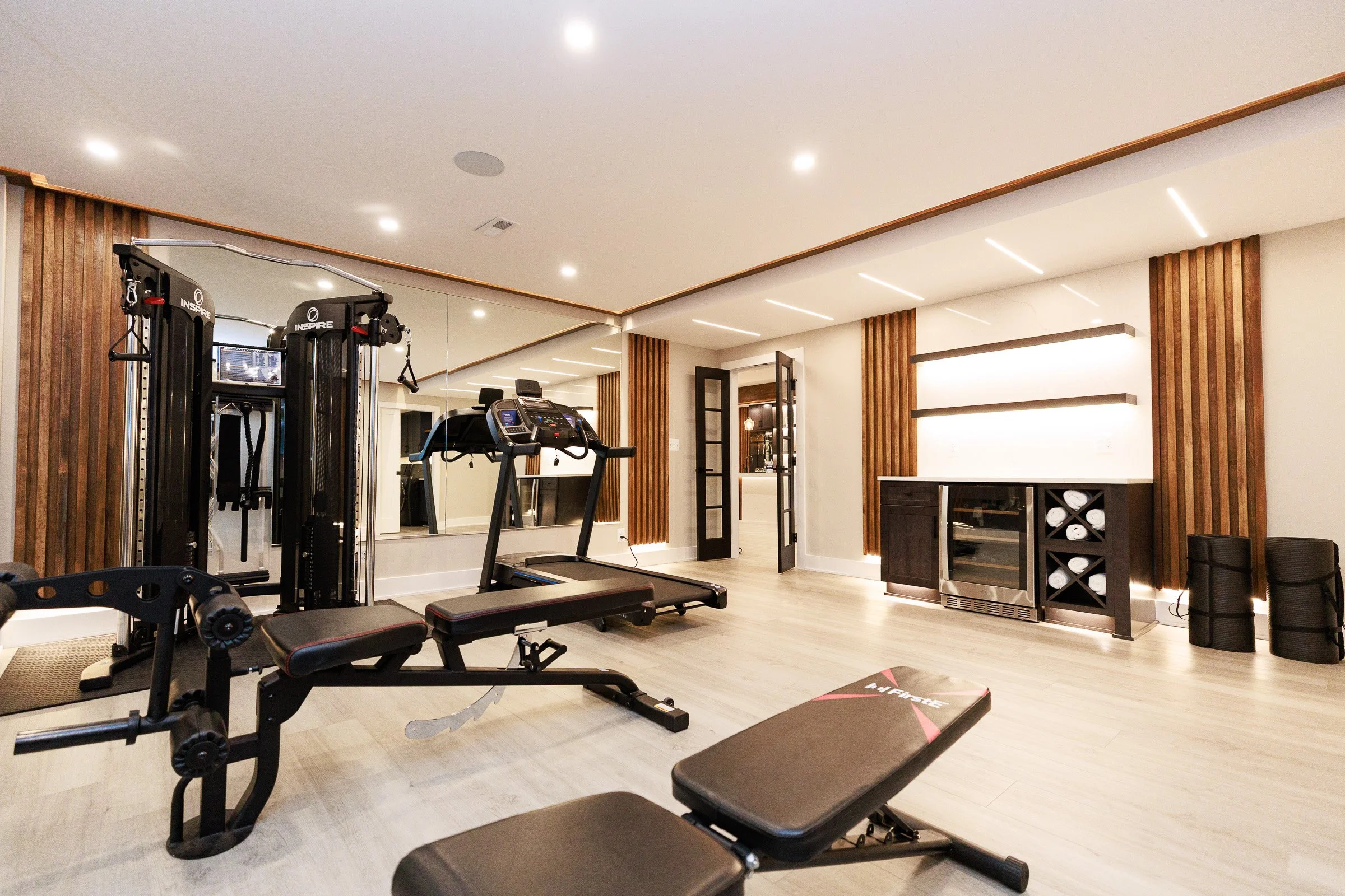 A home gym with workout equipment including a treadmill, weight machine, weight bench, and storage cabinets, decorated with wood paneling and modern lighting.