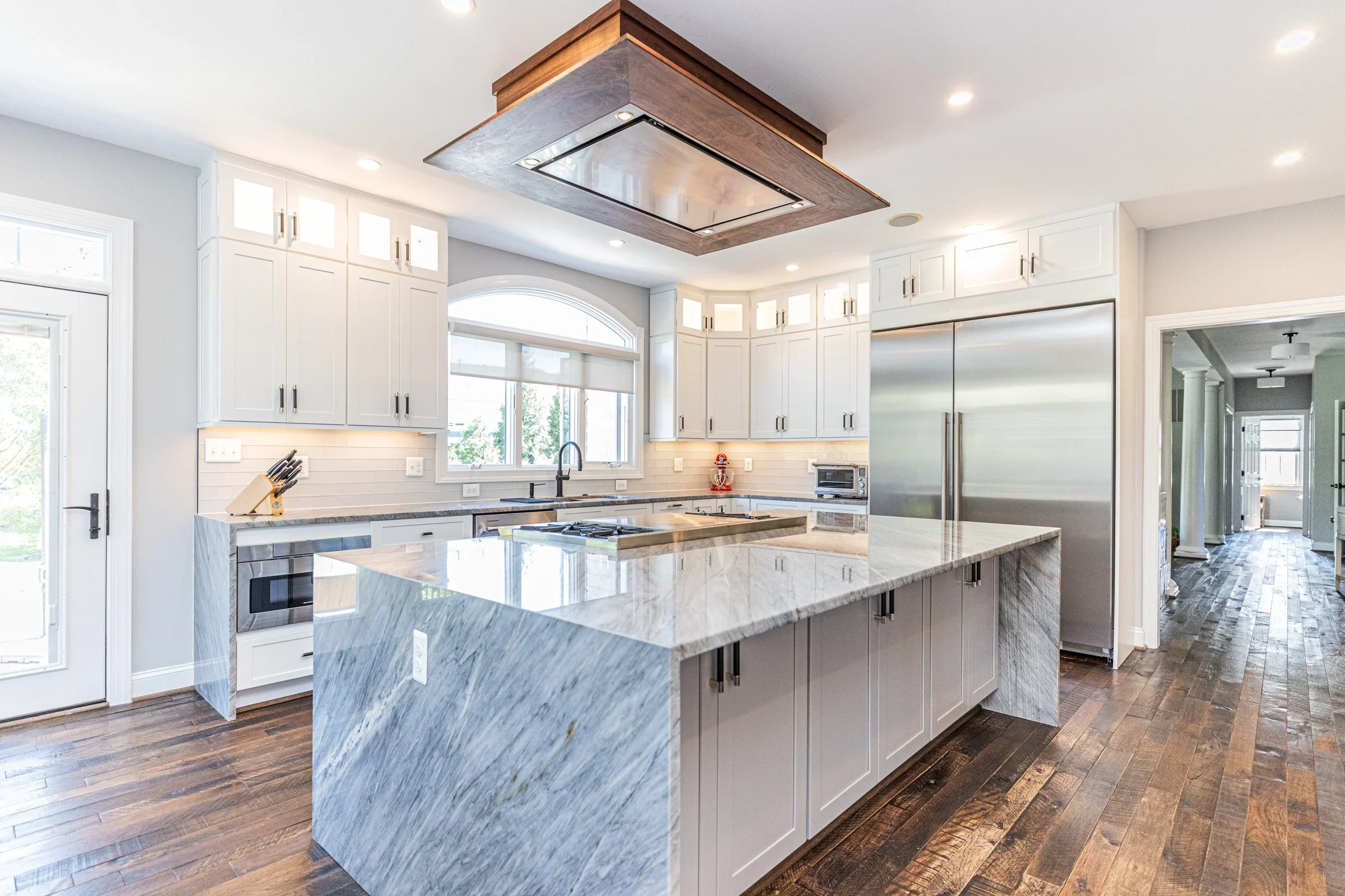 Modern kitchen with white cabinets, marble island, stainless steel refrigerator, microwave, and large window with blinds.