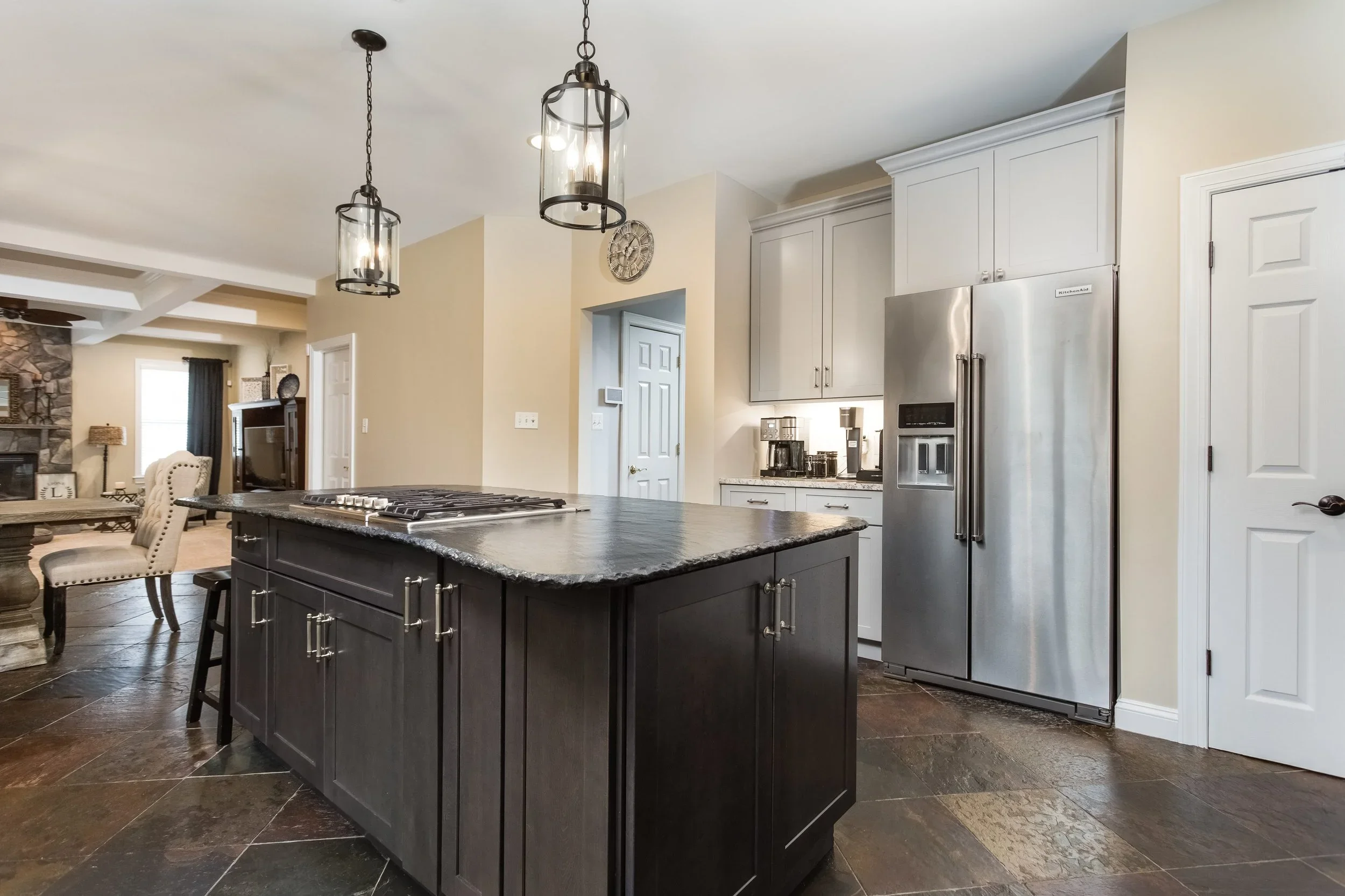 Modern kitchen with a dark island, white cabinets, stainless steel refrigerator, and black pendant lights hanging from the ceiling.