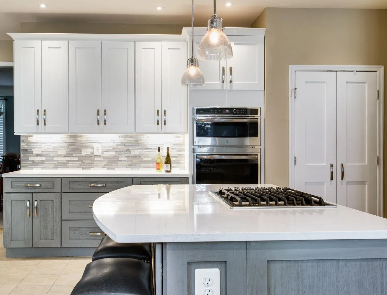 Modern kitchen with white and gray cabinets, marble island, built-in stainless steel oven, and gas cooktop. Pendant lights hang above the counter with bottles placed on the backsplash.