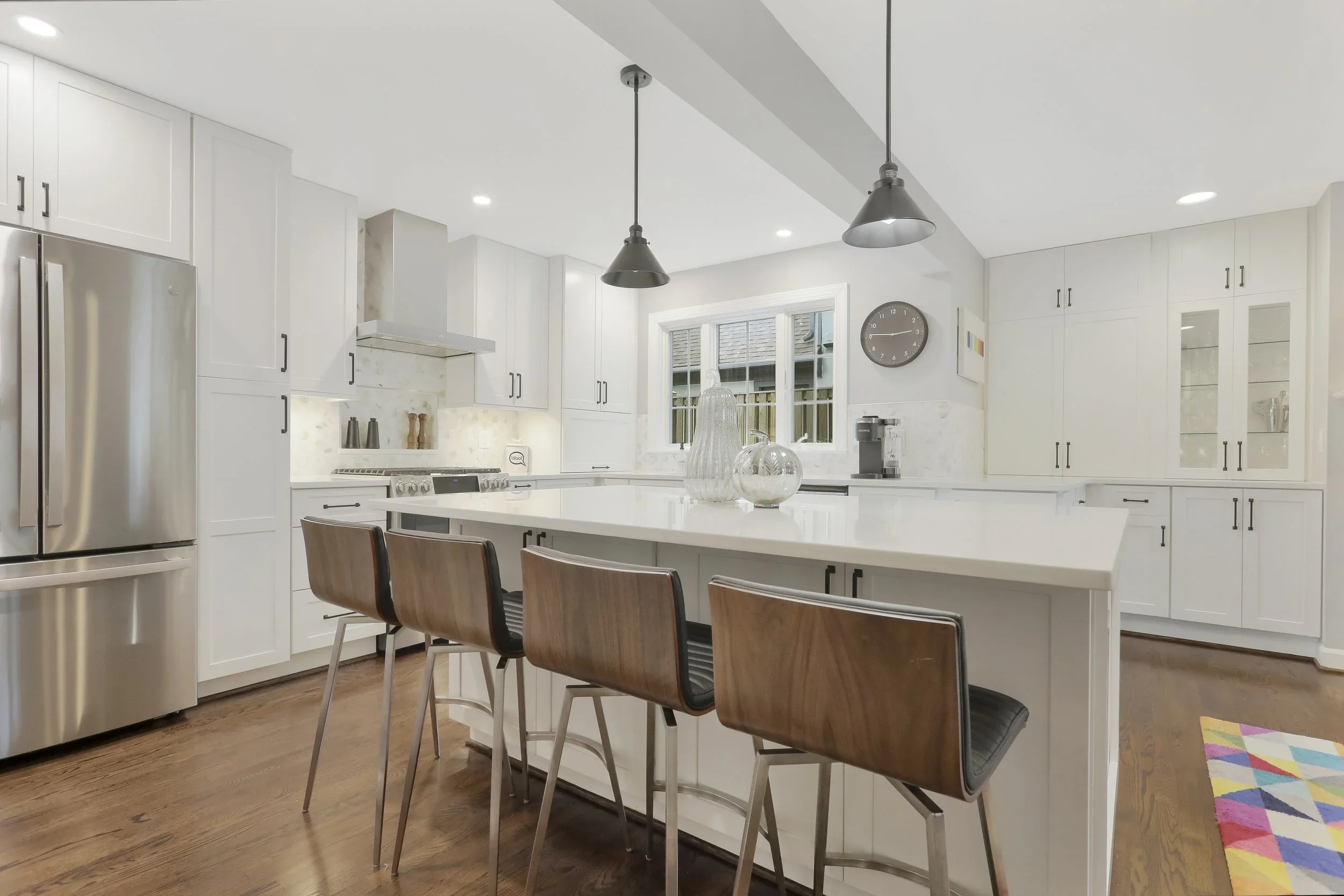 Modern white kitchen with wooden floor, stainless steel refrigerator, white cabinets, central island with four brown chairs, black suspended pendant lights, and a window above the sink.