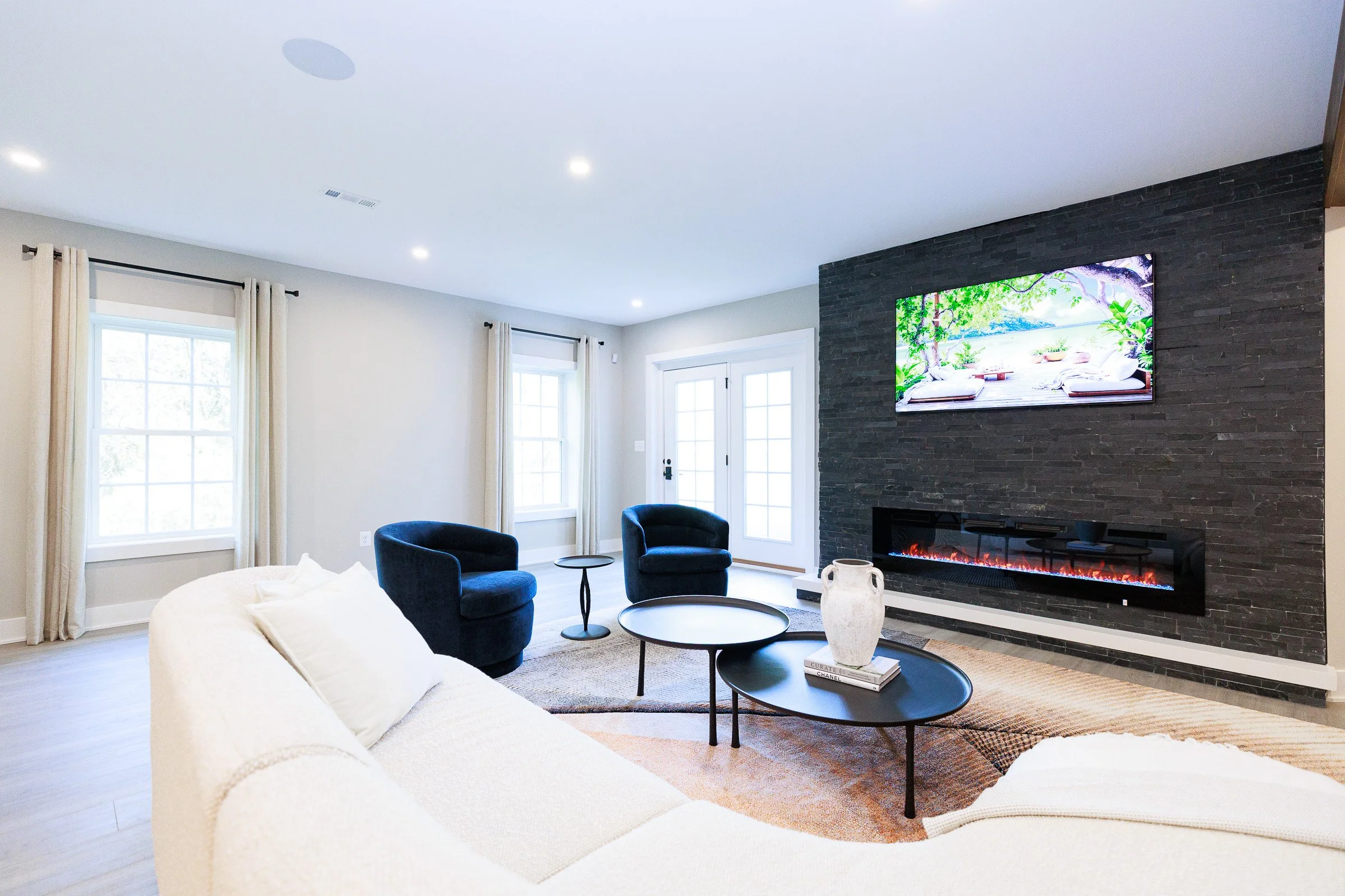 Living room with a white sofa, two dark blue armchairs, a round black coffee table, a fireplace, a wall-mounted TV, large windows with beige curtains, and a door with glass panes.