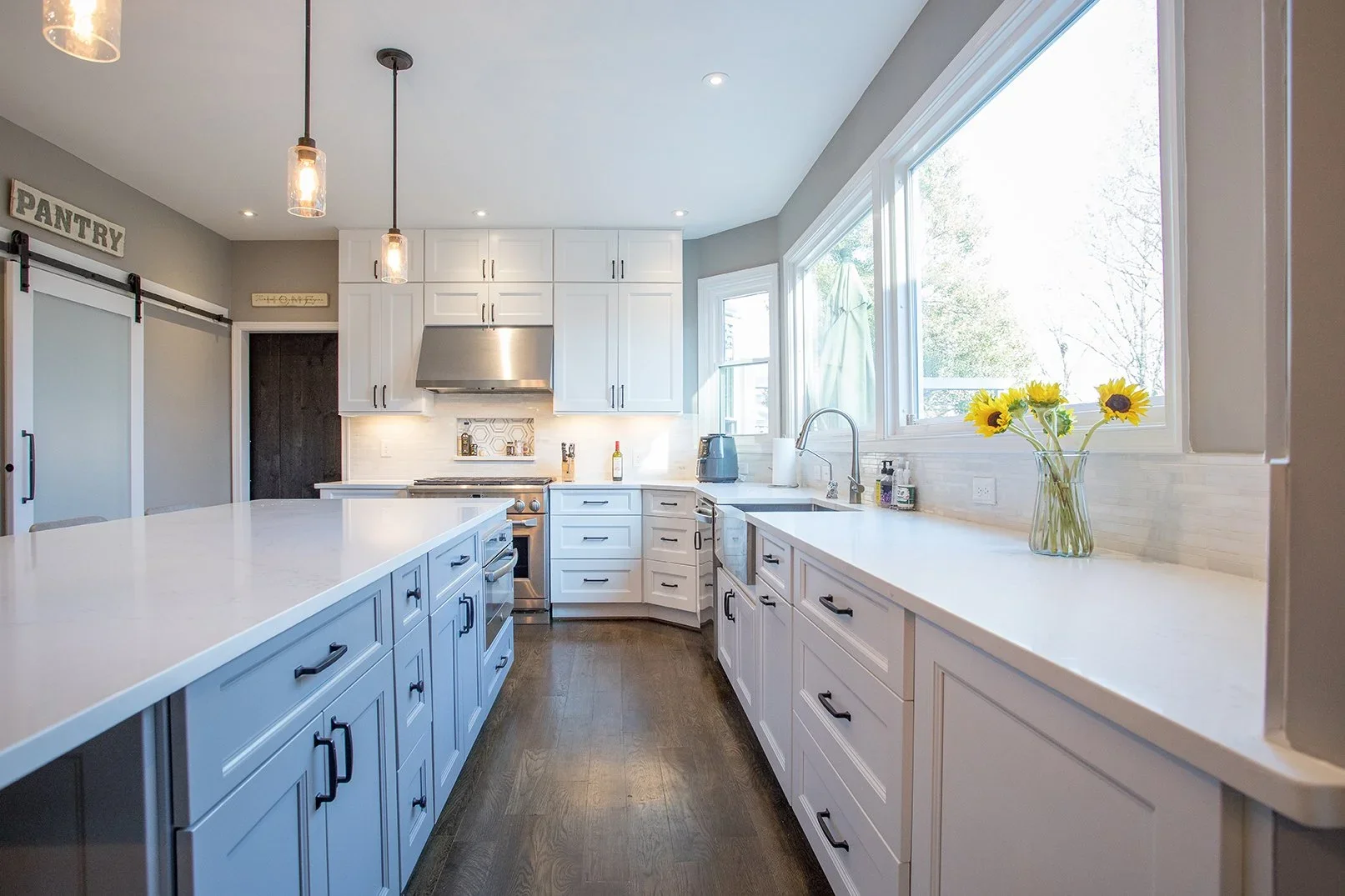 Bright modern kitchen with white cabinets, dark hardwood floors, large windows, and a sunflower vase on the counter.
