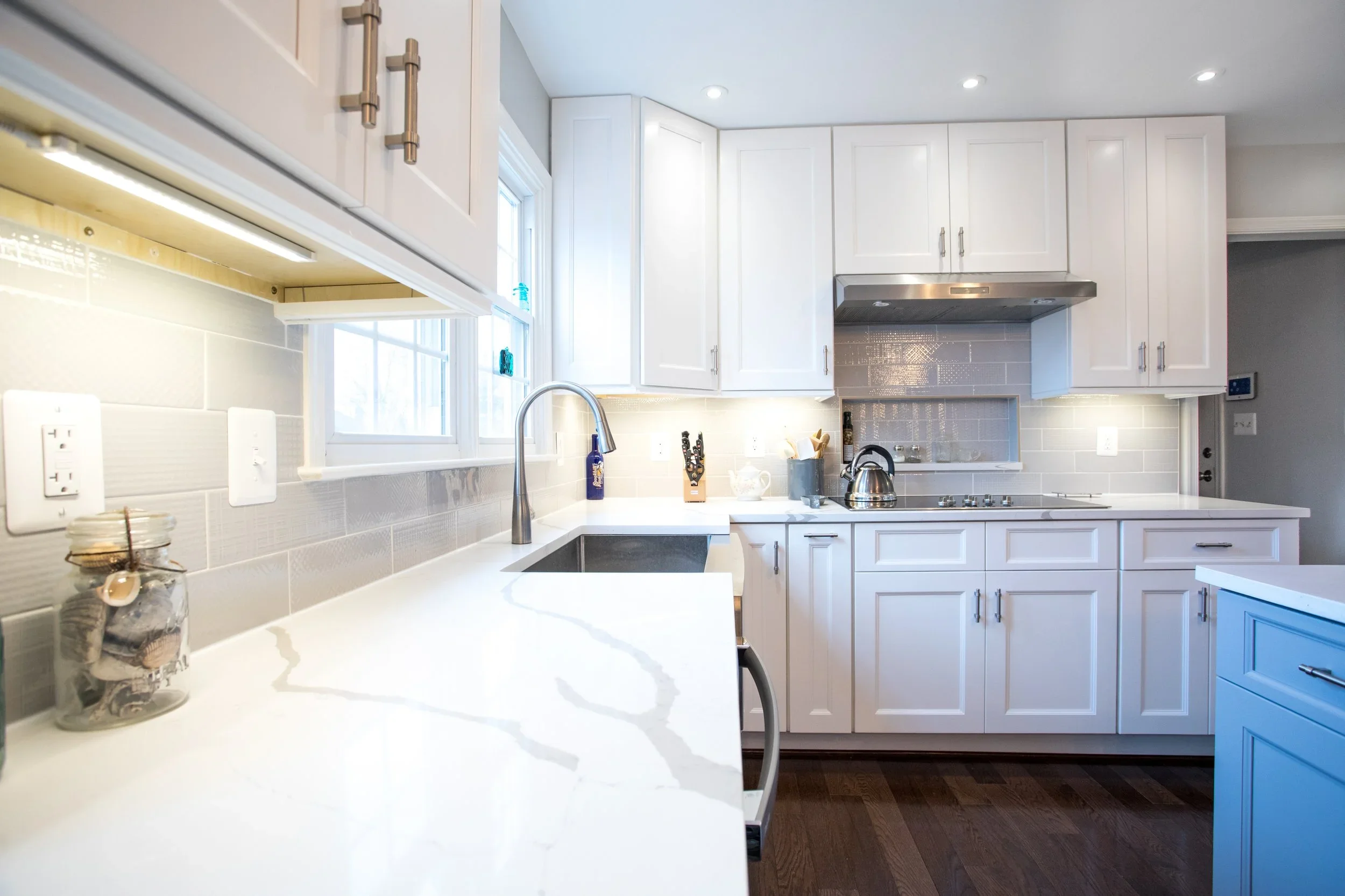 White kitchen with natural light streaming in through windows, featuring a countertop with jars and a jar of tea, white cabinets, stainless steel appliances, and dark wooden floor.