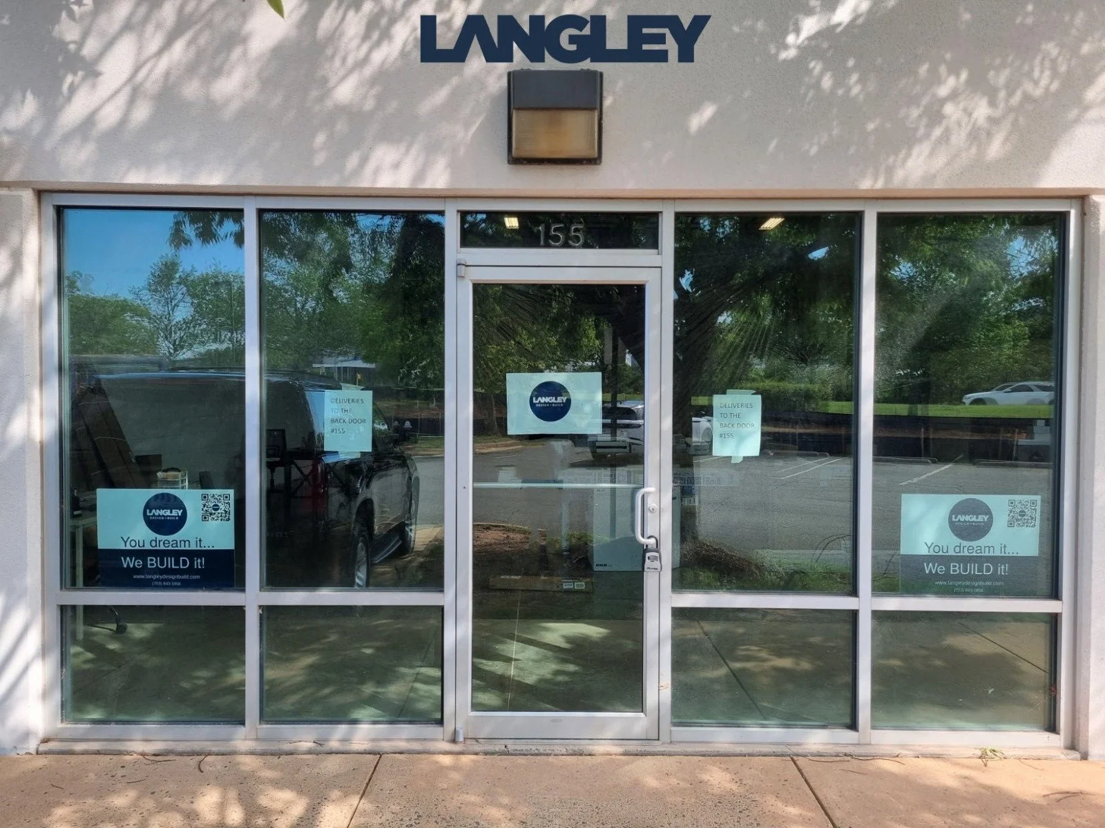 Front view of a commercial building with glass doors and windows showing advertisements for Langley, a construction company. The building is numbered 155 and has signs indicating delivery instructions to the back door.