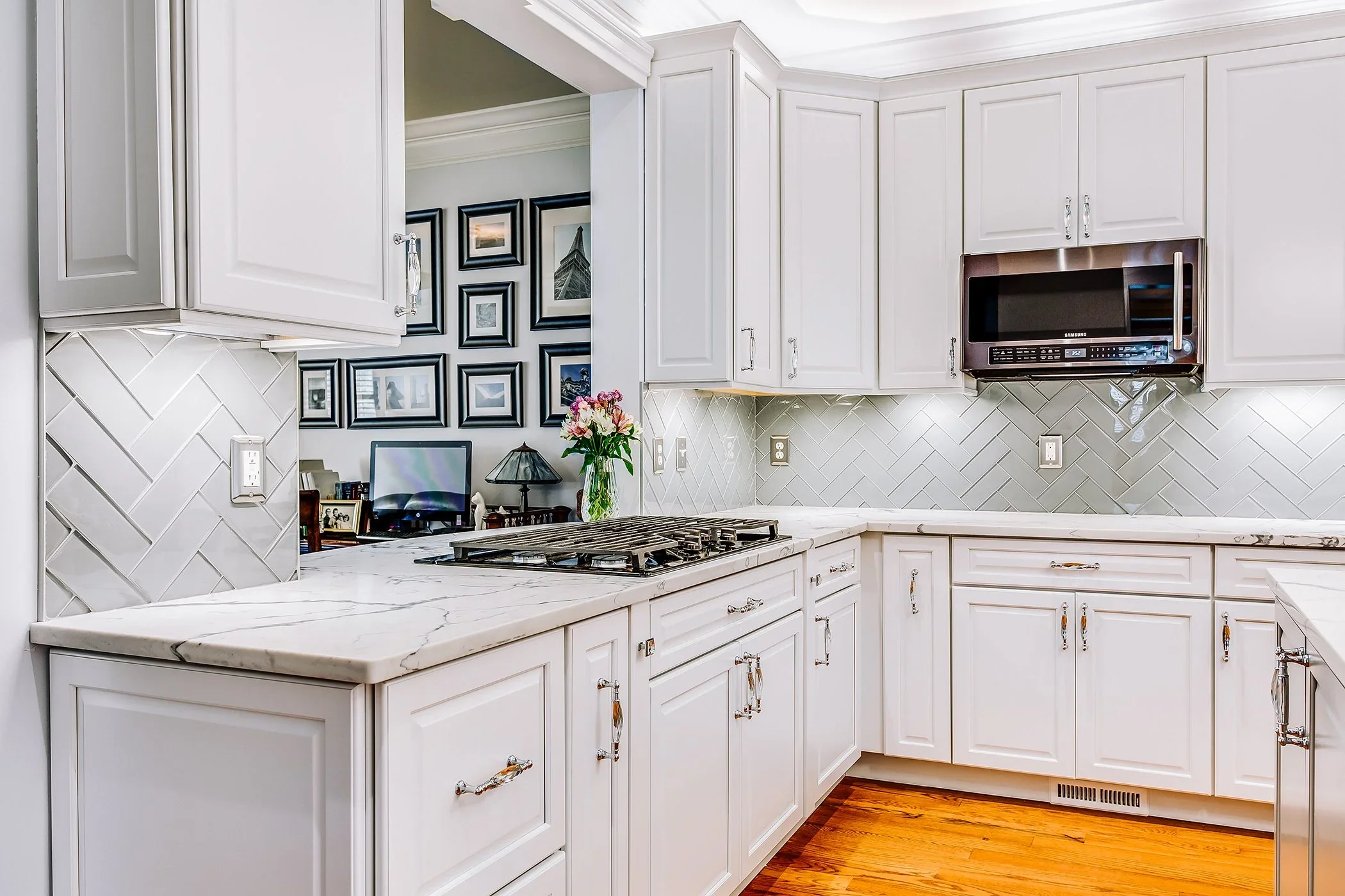 A modern kitchen with white cabinets, a marble countertop, and a backsplash with a herringbone tile pattern. There is a small section of wall with framed photos and a TV mounted on the wall.