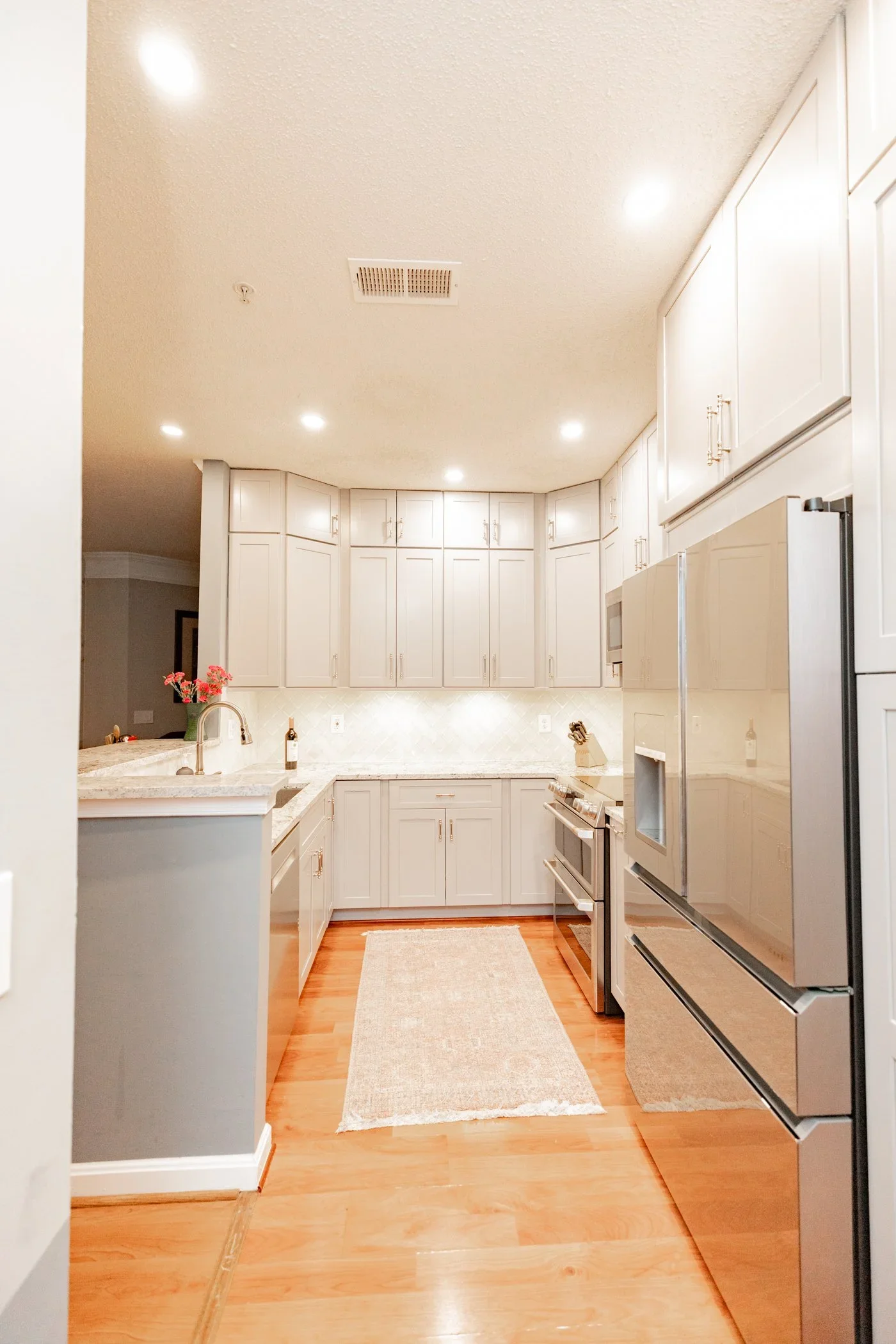 A white kitchen with wooden floors, white cabinetry, stainless steel appliances, and small decorative items on the counter, including a small bouquet of pink flowers in a green vase.