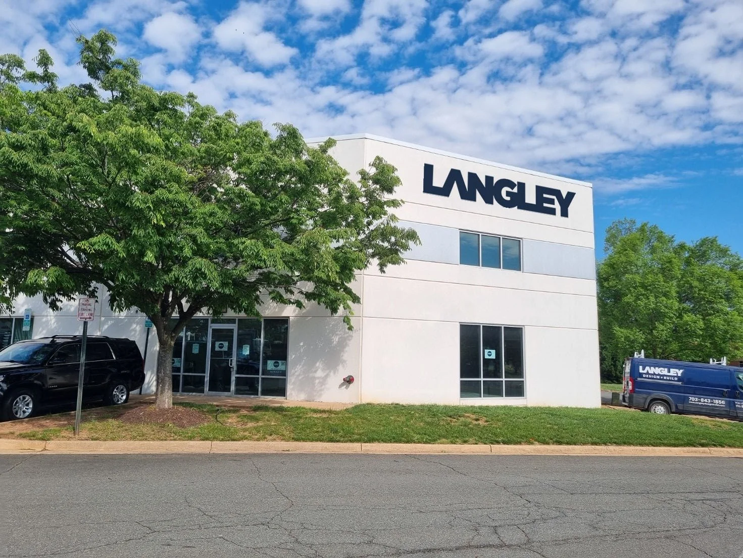 Exterior of a white LANGLEY office building with large sign, parking lot with cars, a green tree in front, and blue sky with clouds.