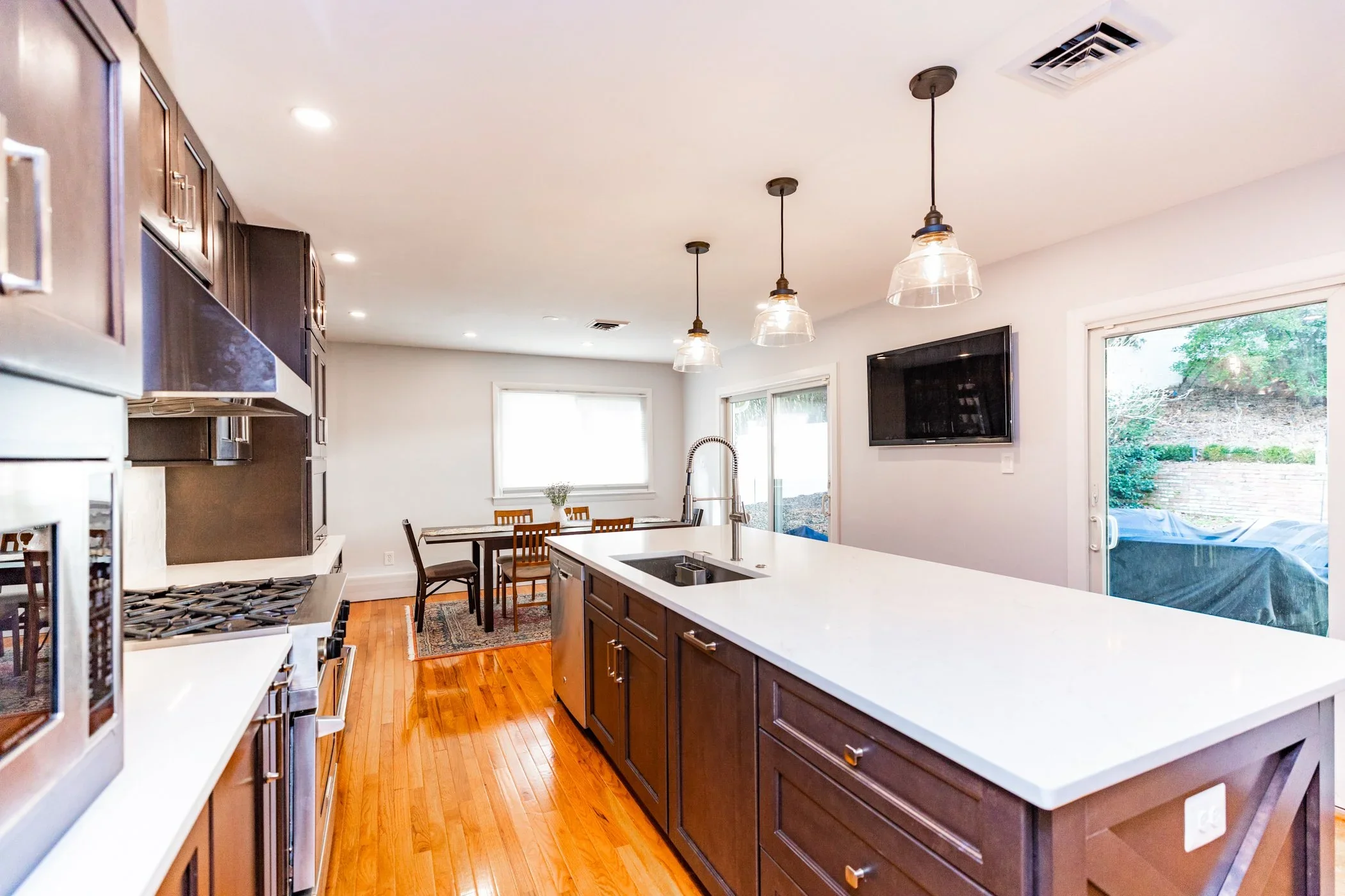Kitchen with dark wood cabinets, white island with sink, stainless steel appliances, hardwood floors, and a dining table with chairs near sliding glass door and window overlooking backyard.
