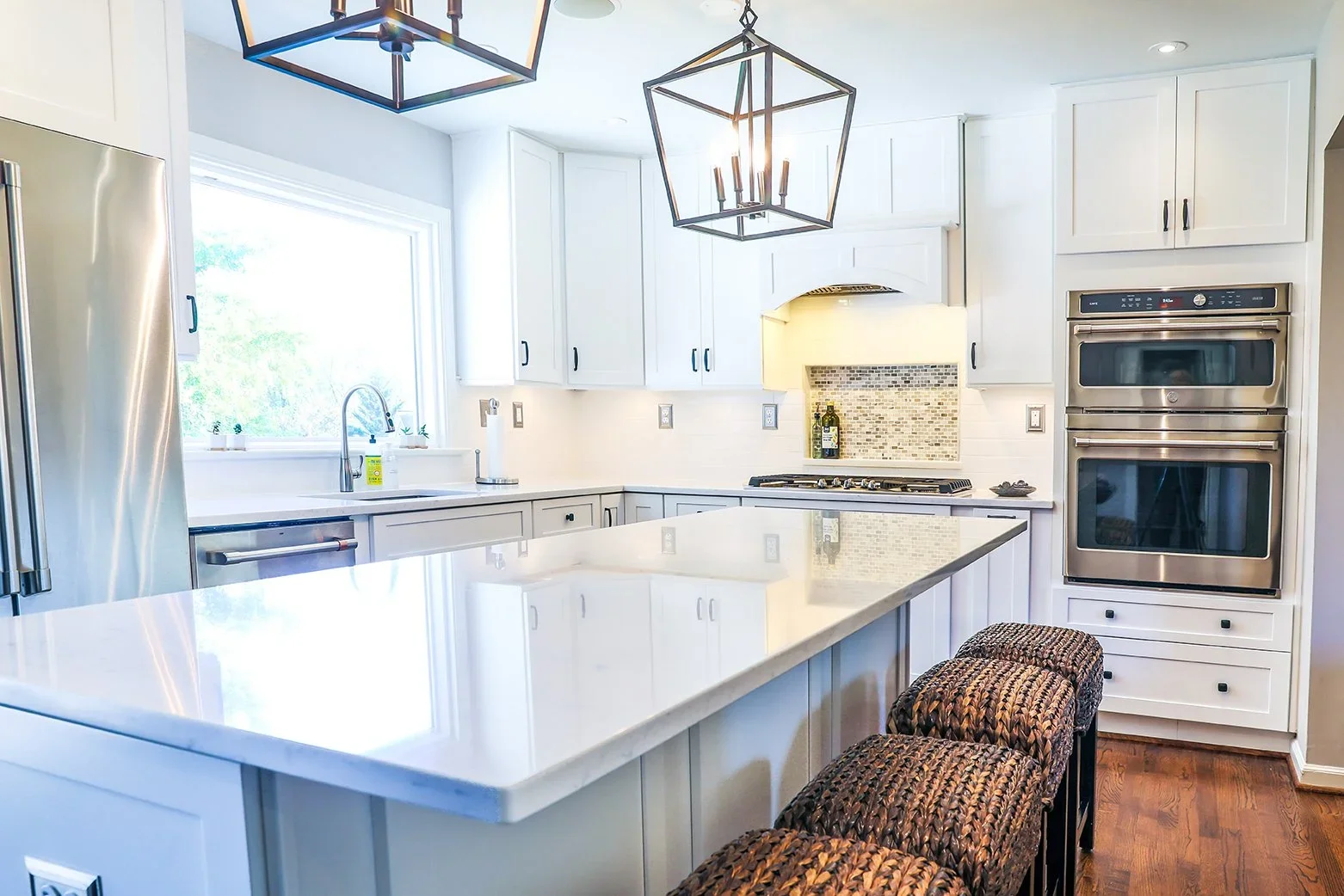 Bright kitchen with white cabinets, marble countertop island, stainless steel refrigerator, double wall oven, gas stove, dual sinks by a large window, and pendant lighting fixtures.